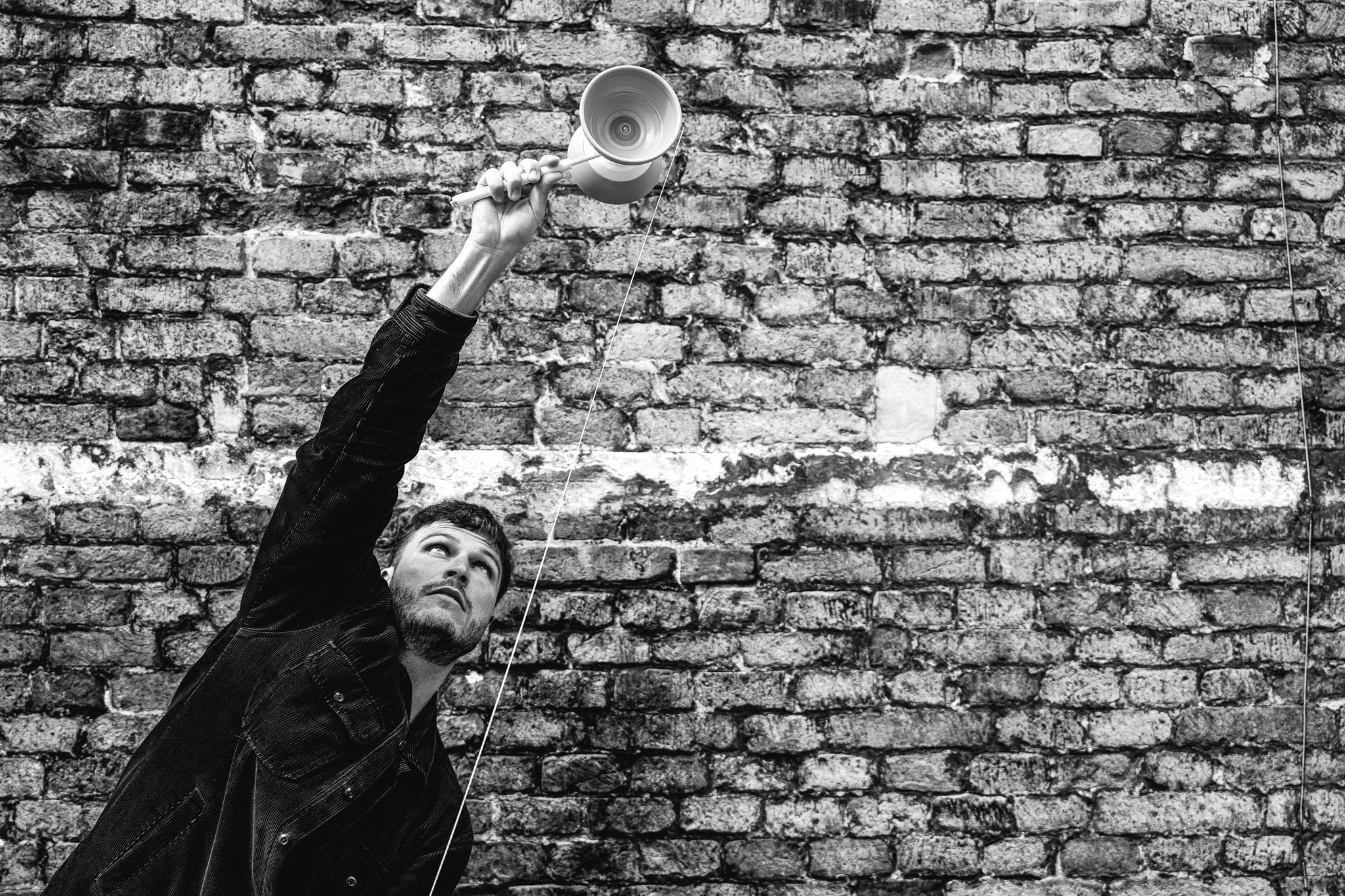 Un homme tient une perruque de style clown au-dessus de sa tête, regardant vers le haut, devant un mur en briques