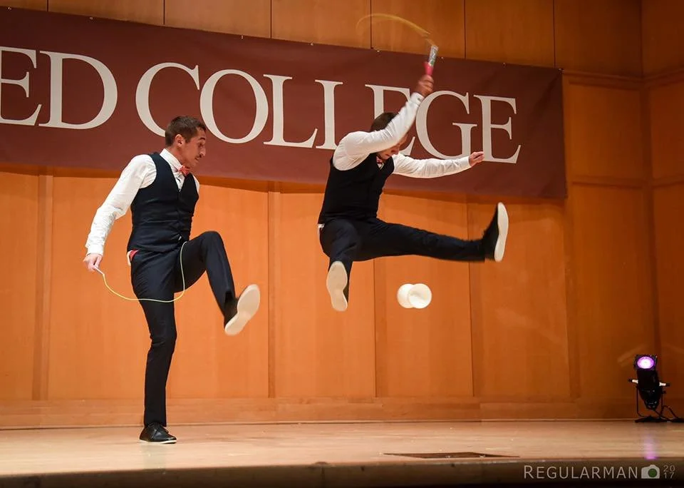 Deux jeunes hommes habillés formellement jouent avec des chapeaux et une corde à sauter sur scène dans une salle de spectacle. Un des deux est en train de sauter en l'air, l'autre est en train de sauter également tout en tenant une corde à sauter.