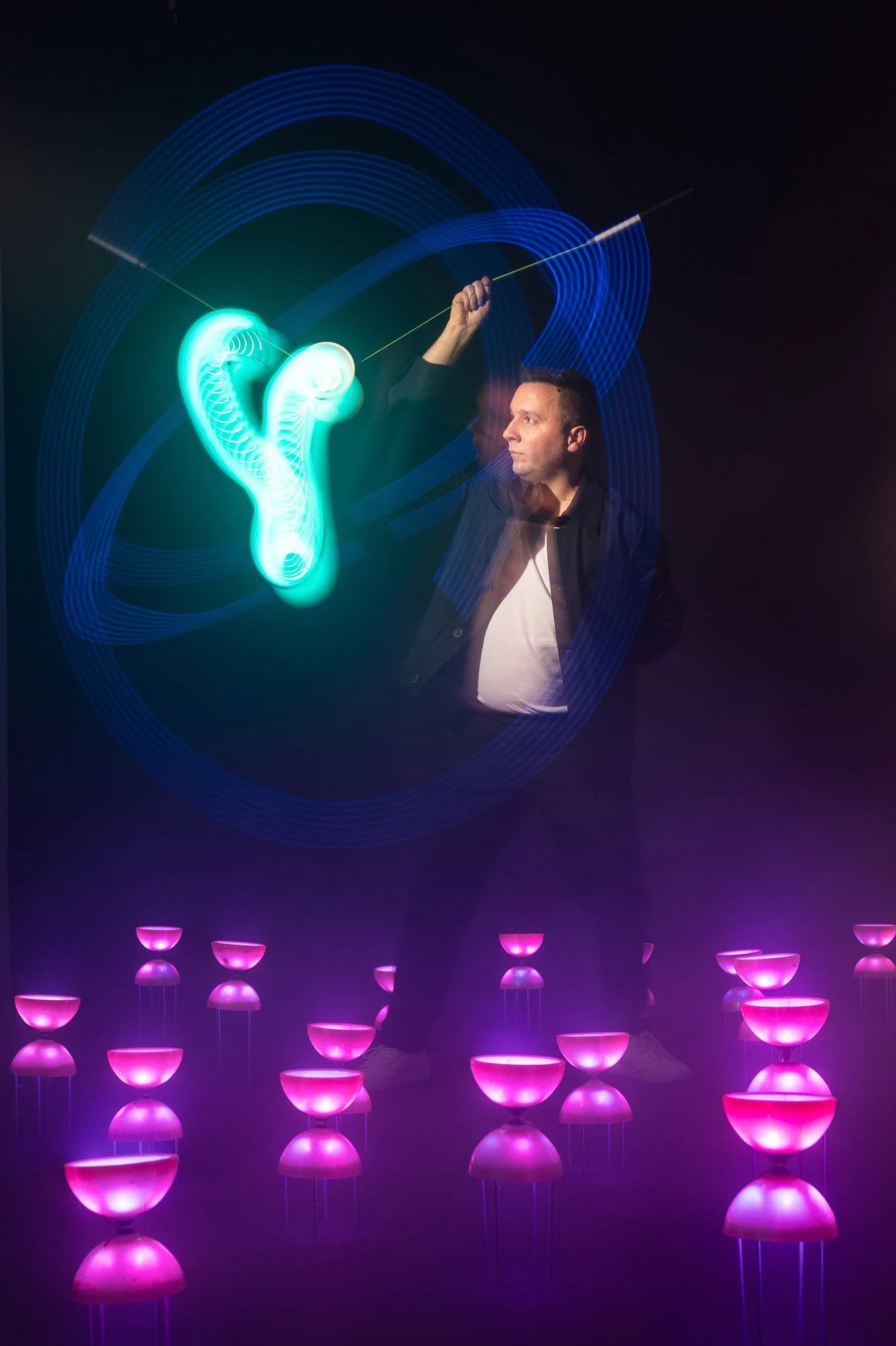 Un homme blanc tenant une baguette ou un stylo, avec des motifs lumineux et des effets de lumière bleue et rose autour de lui, créant une atmosphère futuriste ou artistique.