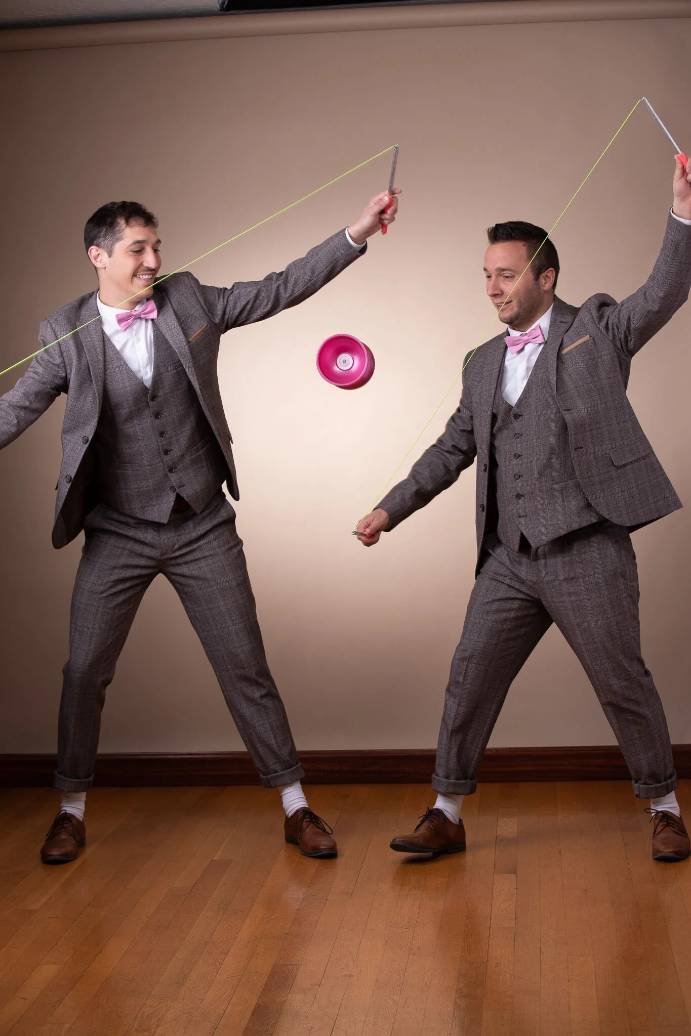 Deux hommes habillés en costumes gris avec nœuds papillon roses, faisant de la jonglerie avec un yo-yo dans une pièce avec un mur beige et un sol en bois.