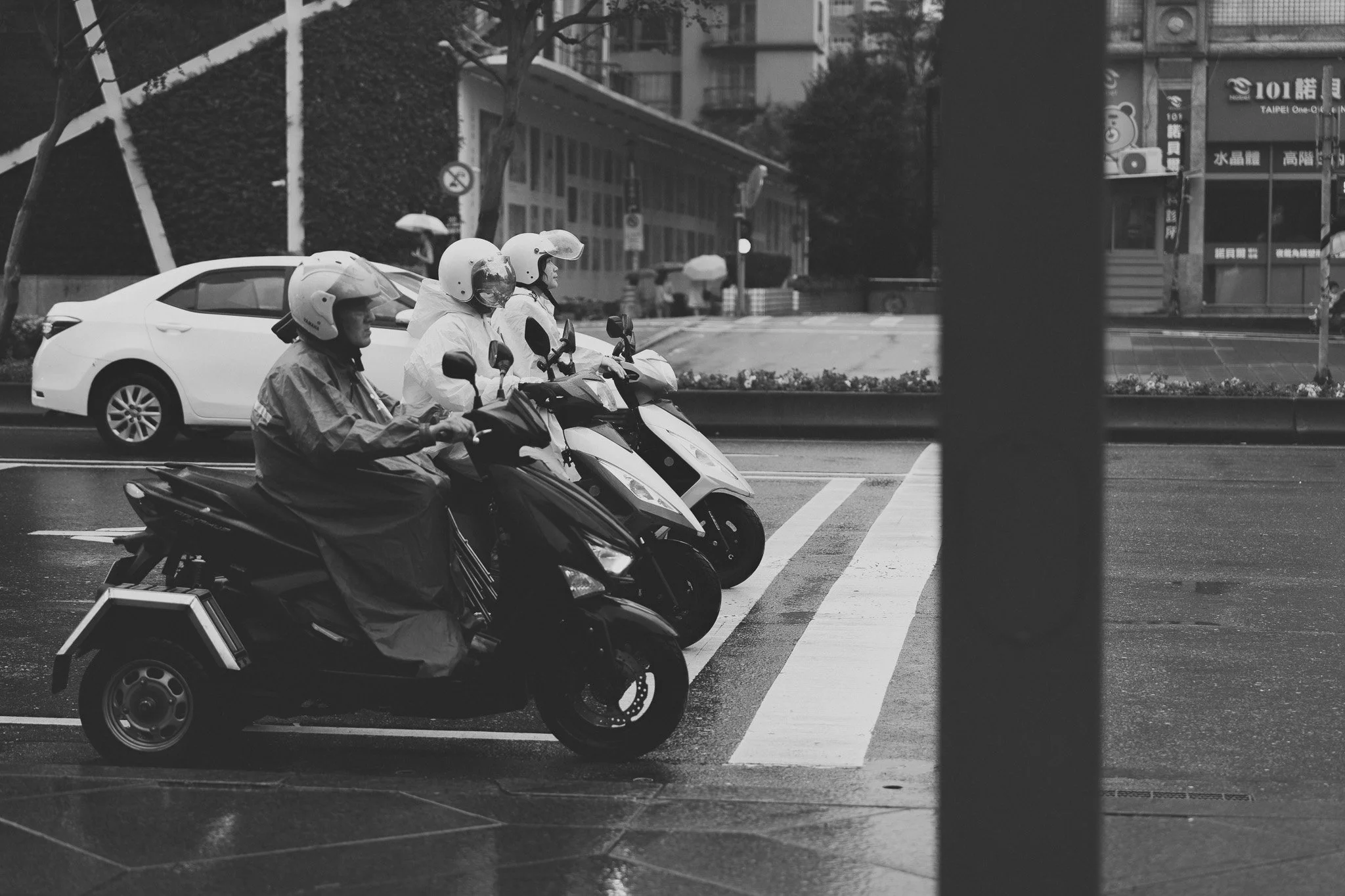 Quatre personnes portant des casques et des imperméables blancs ou gris à scooter, attendent au passage piéton dans une rue urbaine. Il y a une voiture blanche derrière eux, des bâtiments modernes avec des panneaux publicitaires en arrière-plan, et quelques personnes tenant des parapluies.
