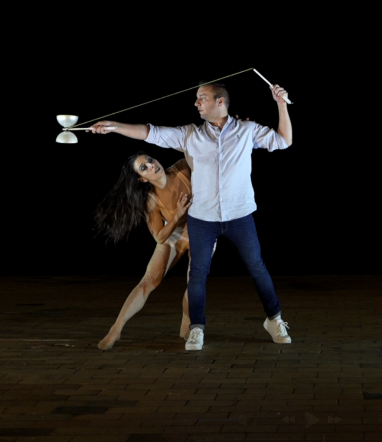 Une personne homme tient une canne d'une main et une femme, vêtue d'une robe de danse, se tient derrière lui dans une pose de danse. Le fond est noir, le sol en bois.