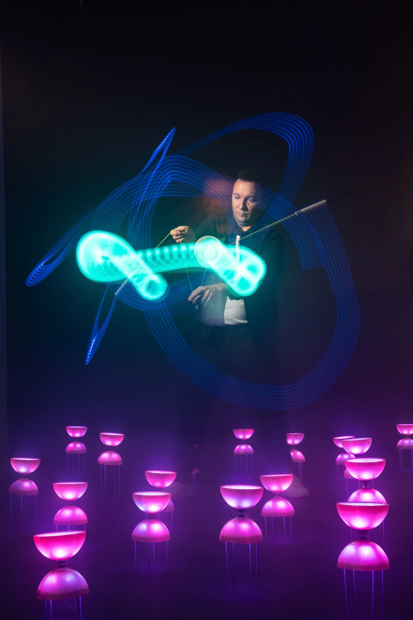 Une personne manipule une expérience de lumière utilisant des objets luminescents et des faisceaux de lumière colorés dans une pièce sombre, avec un plafond rempli de petits points lumineux et des étages de bols lumineux en dessous.
