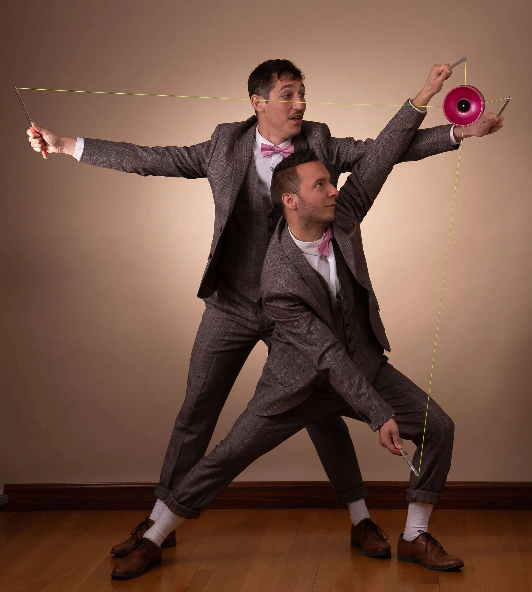 Deux hommes en costume gris avec chemise blanche, l'un portant un nœud papillon rose, dans une pose artistique avec des baguettes et de la ficelle jaune, dans une pièce avec un mur beige et un plancher en bois.