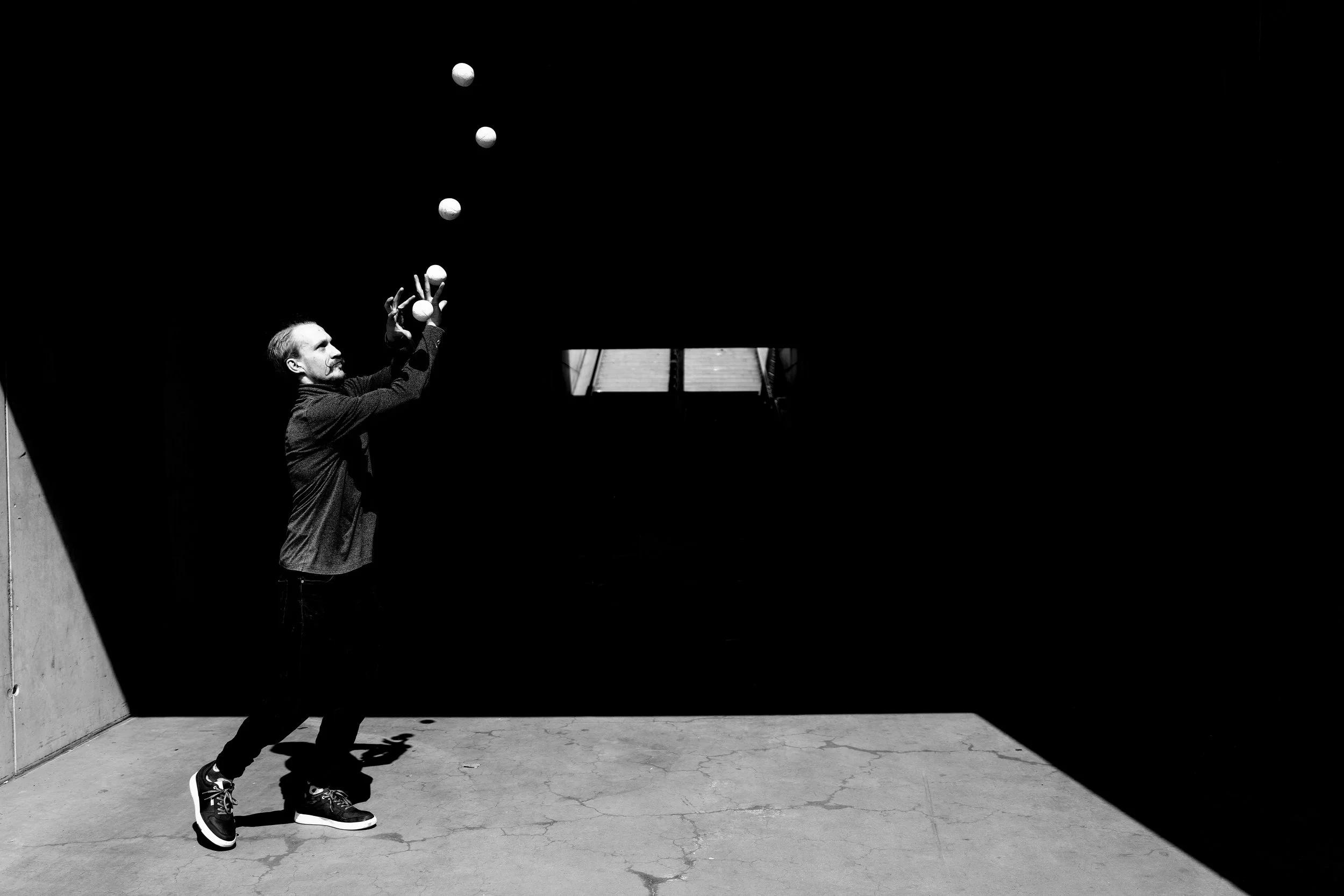 Un homme jongle avec des balles dans un espace sombre, avec une lumière venant d'une ouverture en haut.