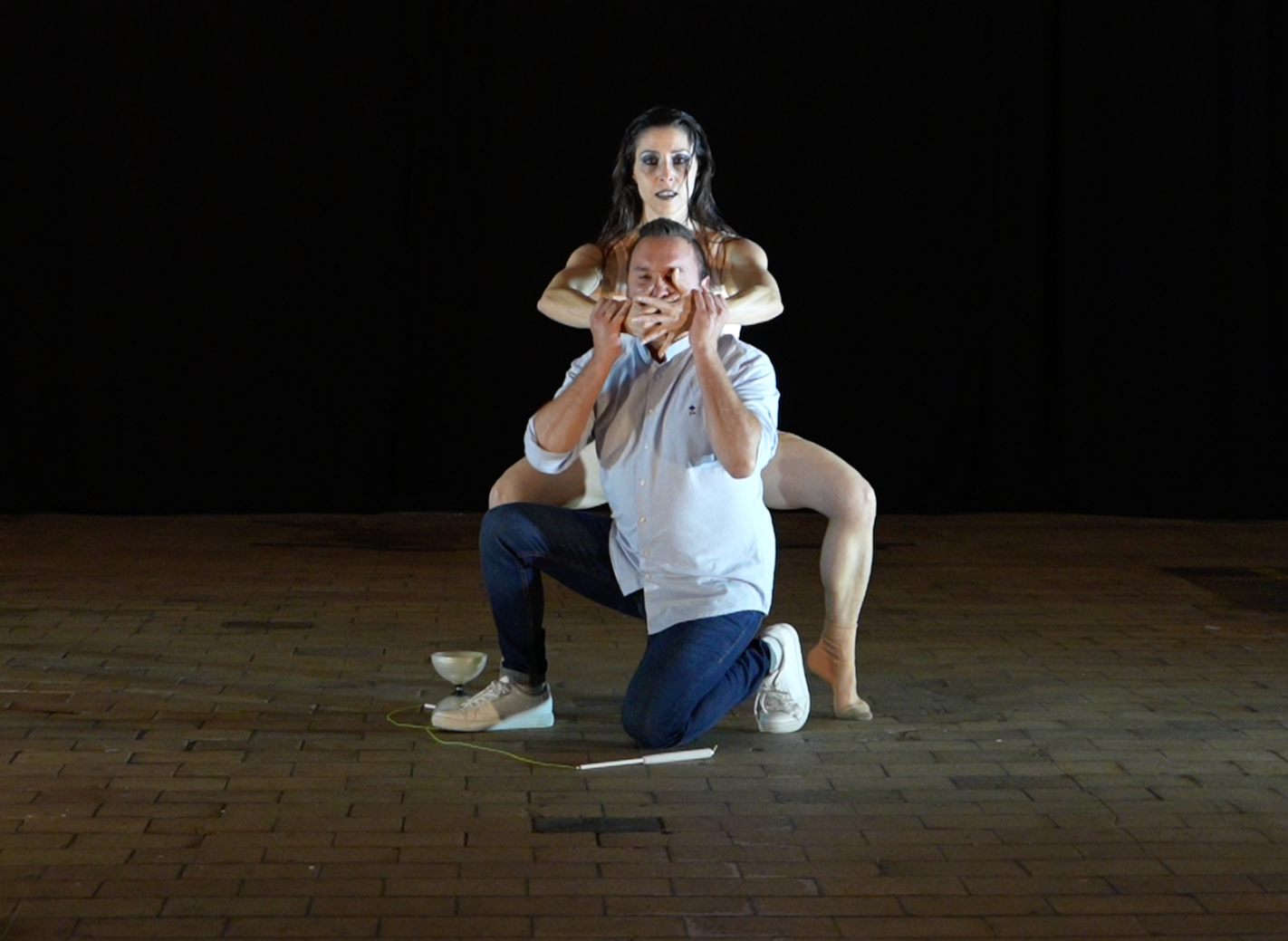 Une performance artistique avec trois personnes. Une femme au centre, en chair et en os, avec une expression sérieuse. Elle est derrière un homme à genoux, qui porte une chemise blanche et des jeans, tenant un morceau de bois près de sa bouche. Une sculpture ressemblant à une femme est placée derrière eux, avec ses bras autour de la tête de l'homme, créant une composition théâtrale sur scène, avec un fond noir et un sol en bois.