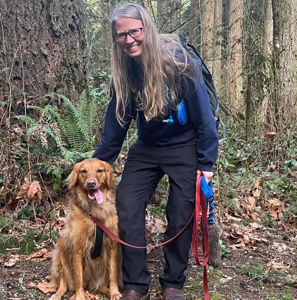 A woman with glasses and long hair smiling while kneeling with her dog in a wooded area.
