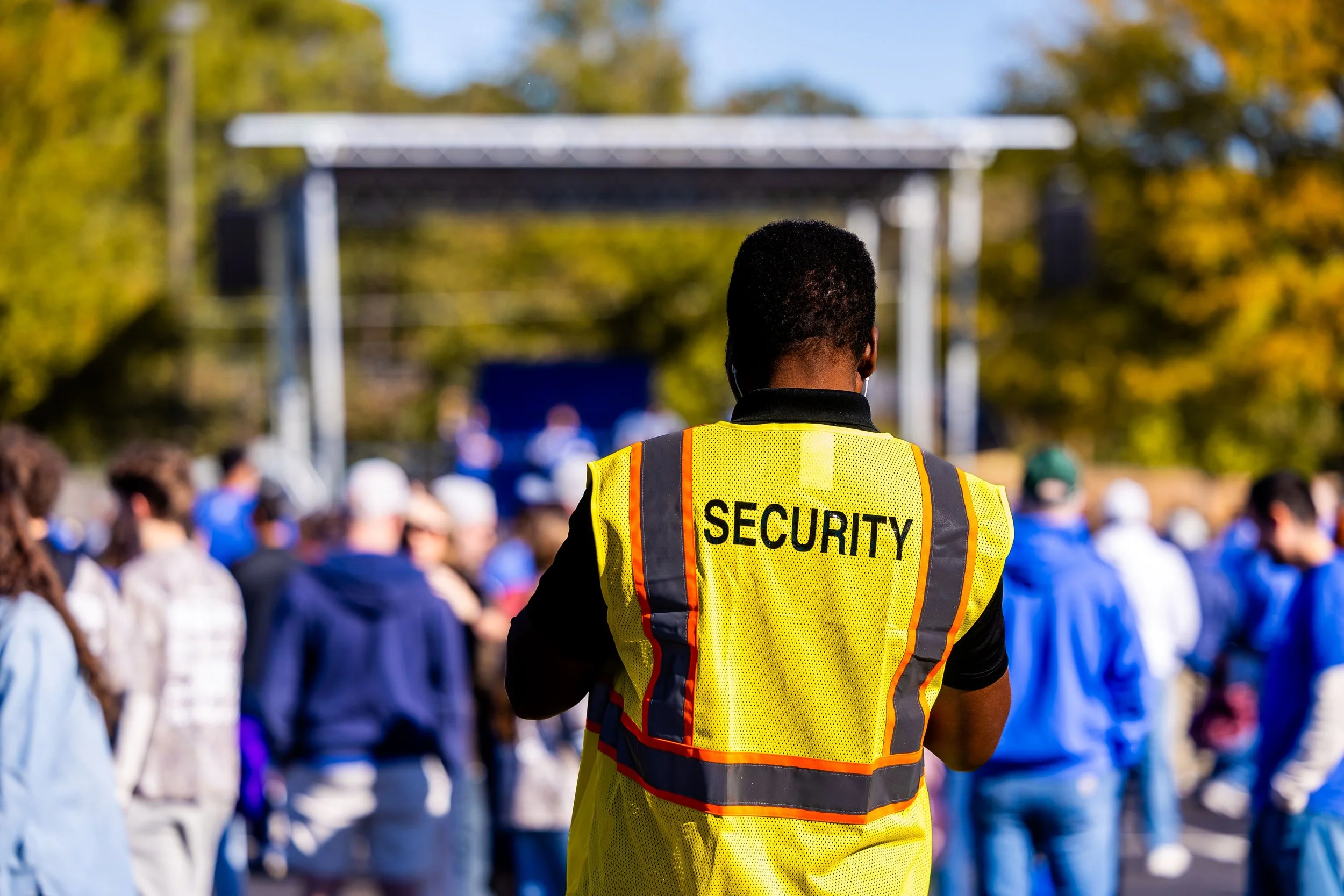 Security guard wearing a high-visibility vest with 'SECURITY' written on the back at a crowded outdoor event with many people and trees in the background.