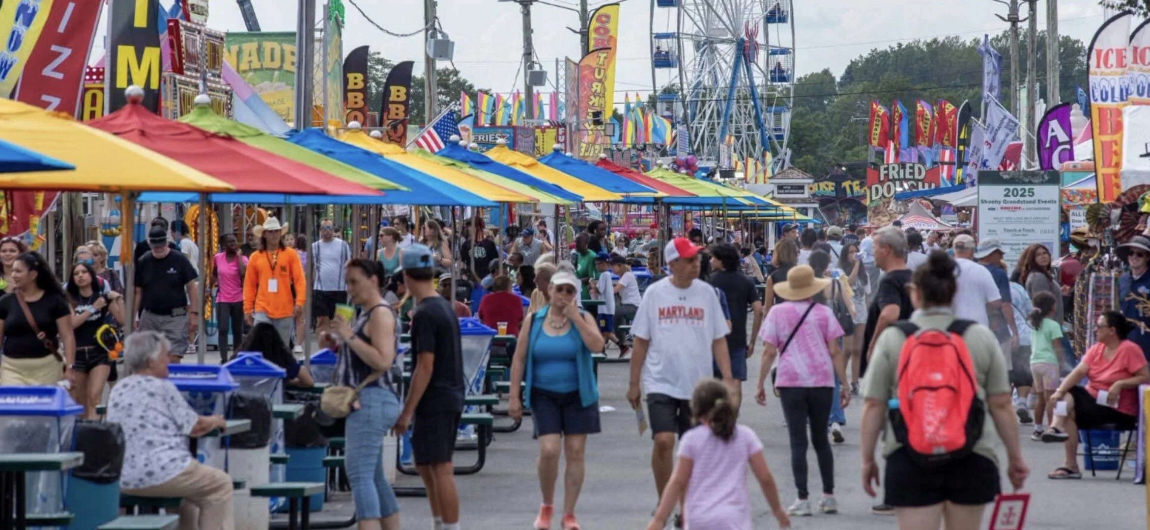 A crowded fairground with colorful tents, rides, and flags, with many people walking and sitting, some eating and playing games.