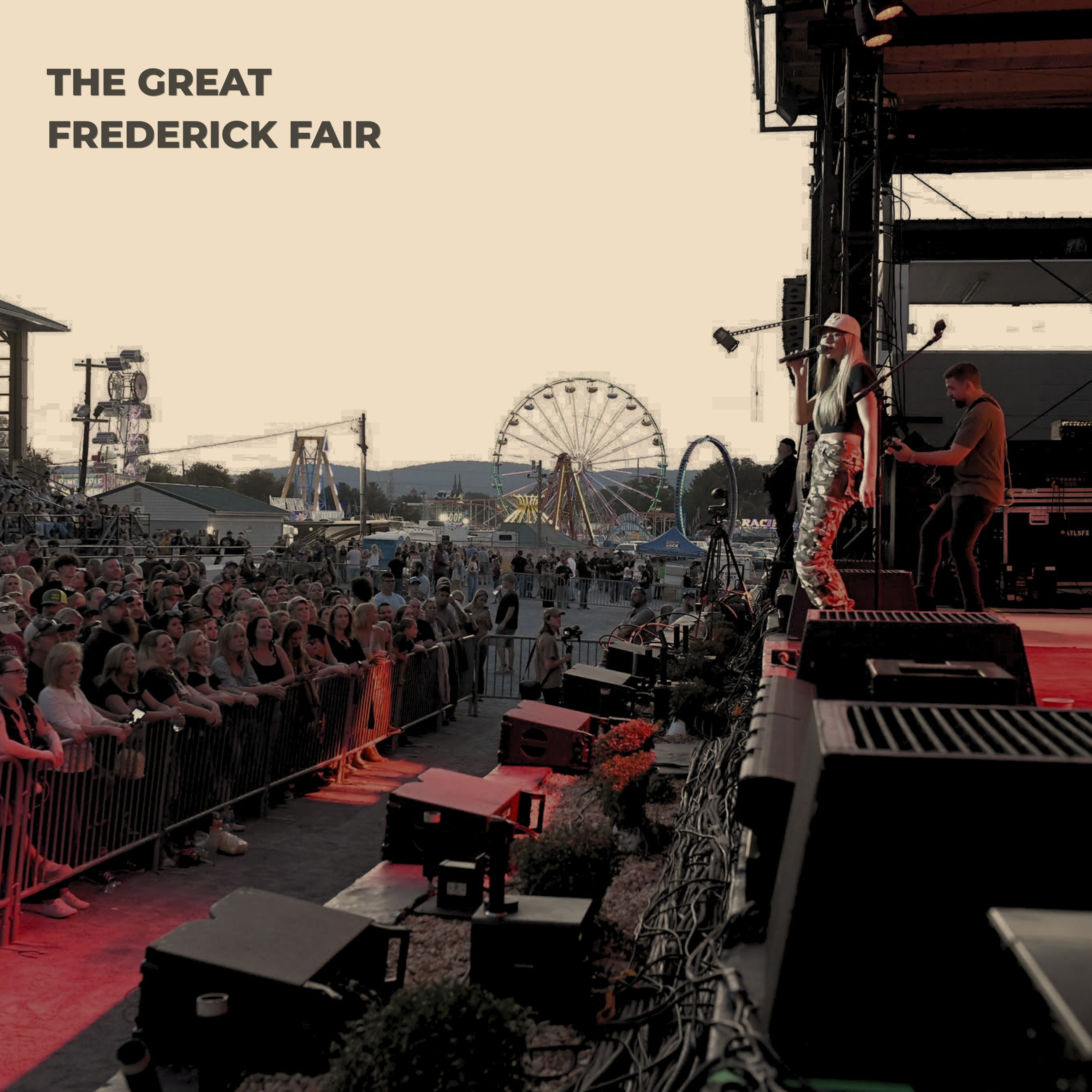Stage at Frederick Fair with a performer singing into a microphone, audience seated in front, amusement rides including a Ferris wheel in the background, during evening.