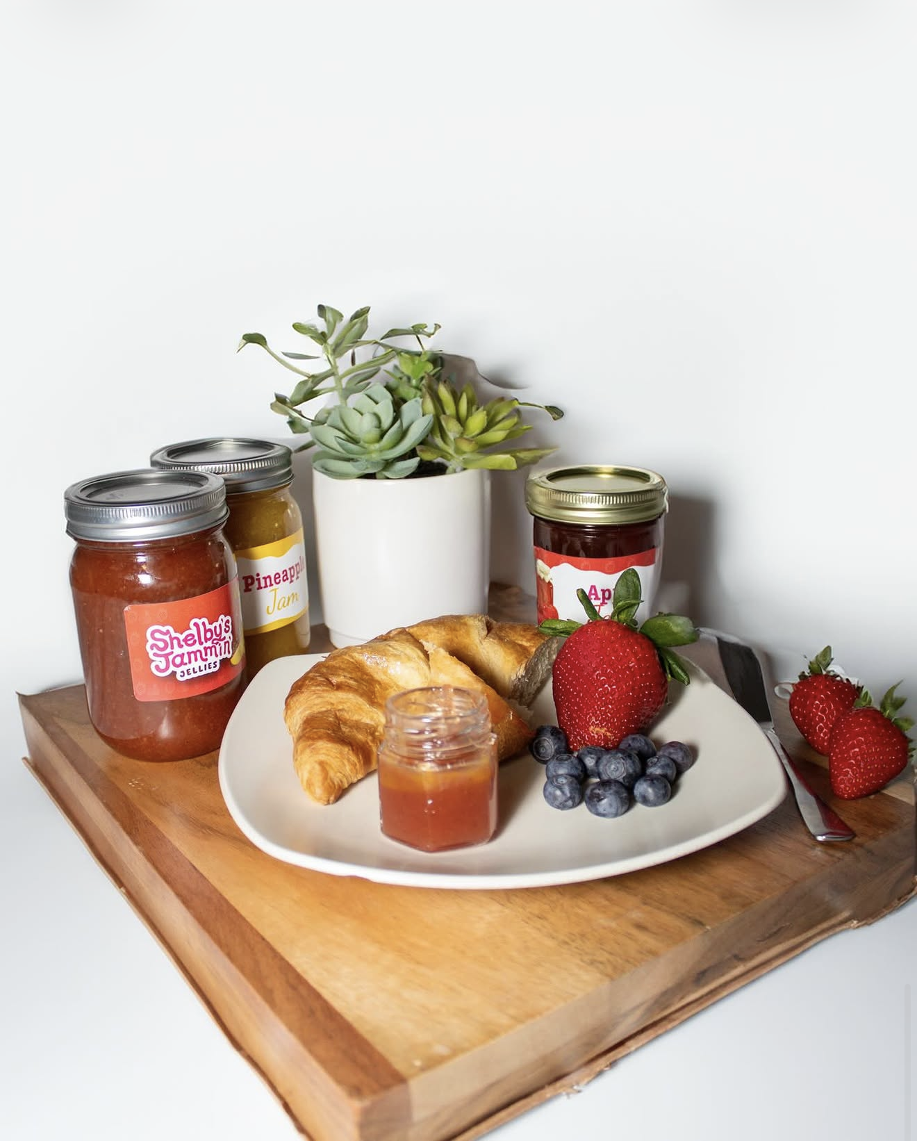 A breakfast scene on a wooden tray with a croissant, fresh strawberries, blueberries, a jar of honey, and jars of jelly and jam, with a small jar of honey in front, and a white plate and knife. In the background, there are three jars of jam and a white pot with green succulent plants against a plain white wall.