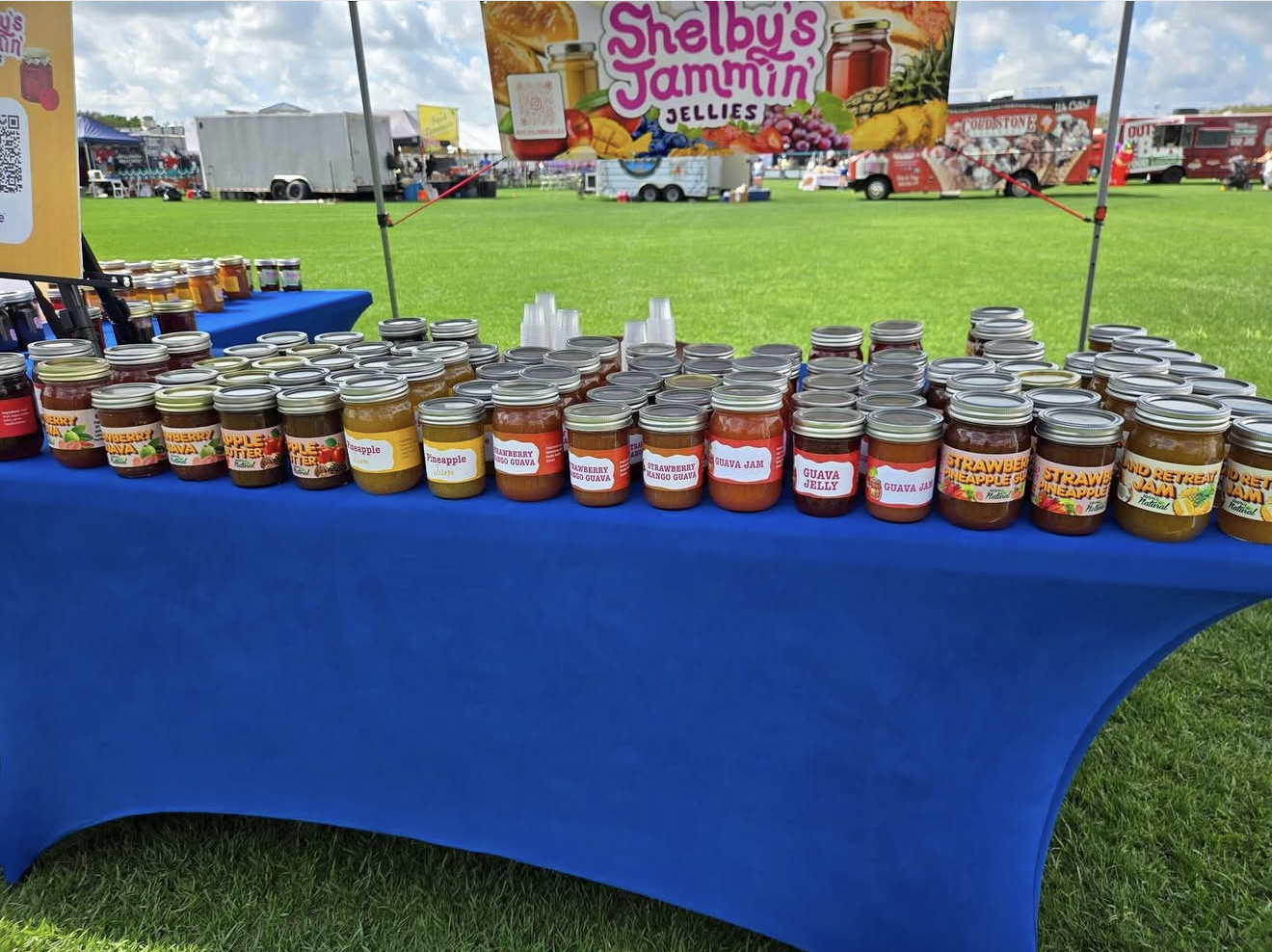 Table covered in jars of jelly and jam at an outdoor fair, with a sign advertising Shelby's Jammin Jellies and a green grassy field in the background.