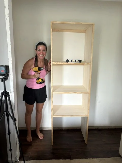 A woman in a pink tank top and black shorts smiling and holding a yellow power drill stands next to a tall wooden shelving unit under construction in a room with hardwood floors and a plain wall.