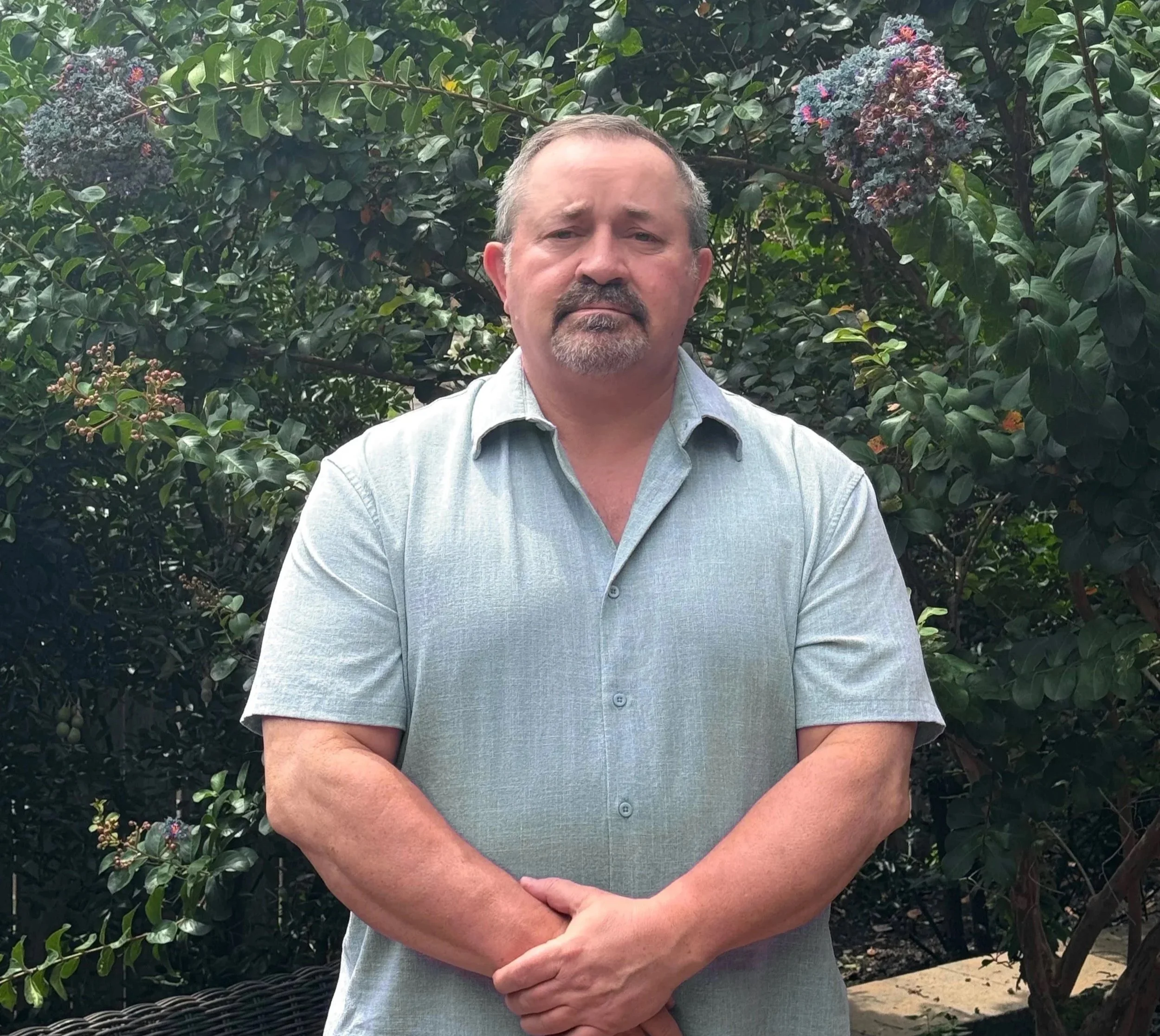 A middle-aged man with short hair and a goatee stands outdoors in front of green foliage with purple flowers, wearing a light blue button-up shirt, crossing his arms.