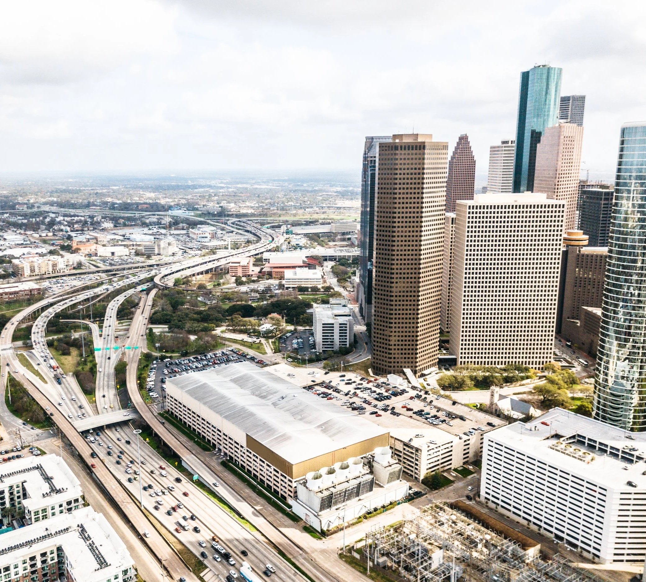 A cityscape with tall buildings, highways, and parking lots, under a partly cloudy sky.