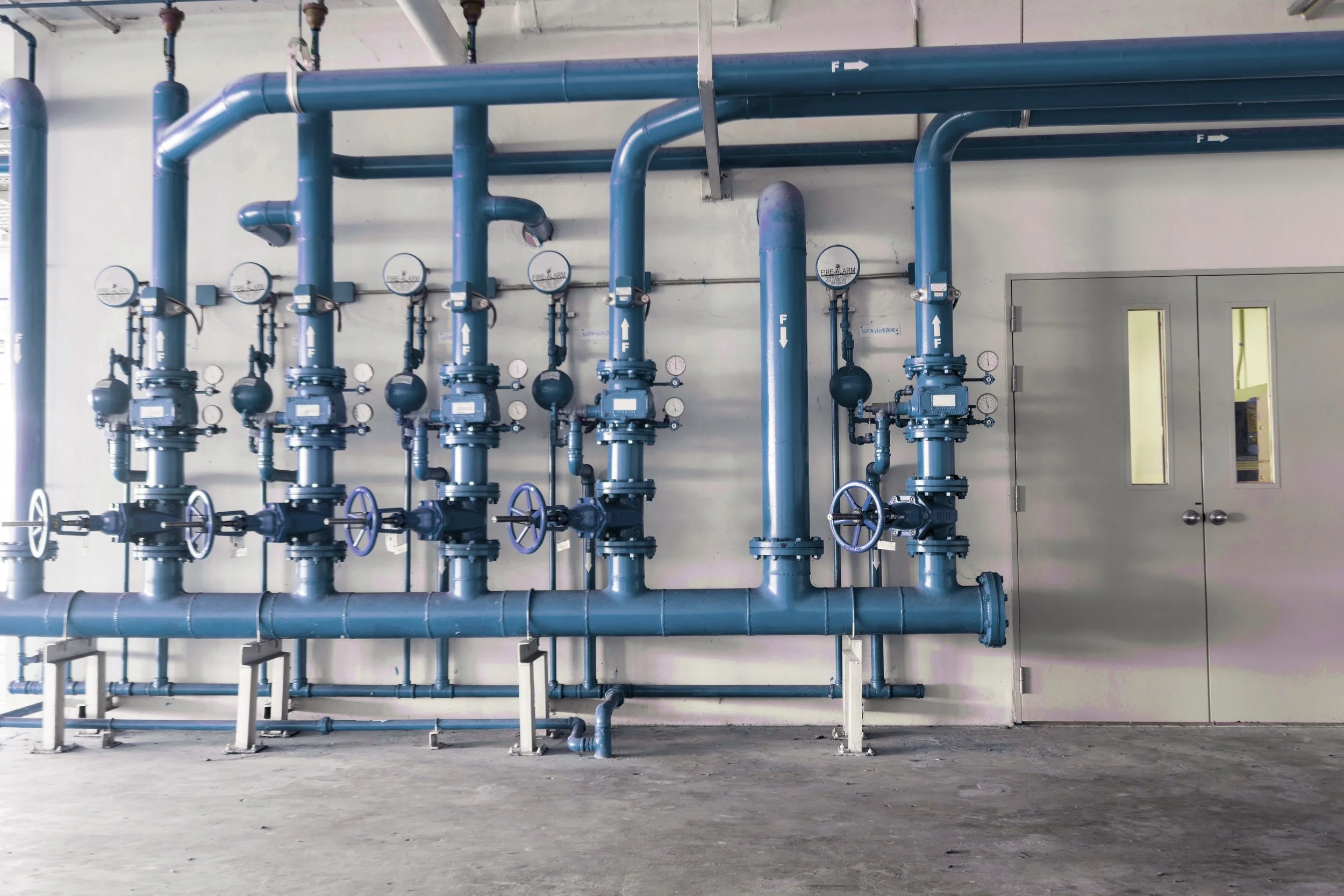Industrial room with blue pipes, gauges, and valves on a white wall and a closed gray door.
