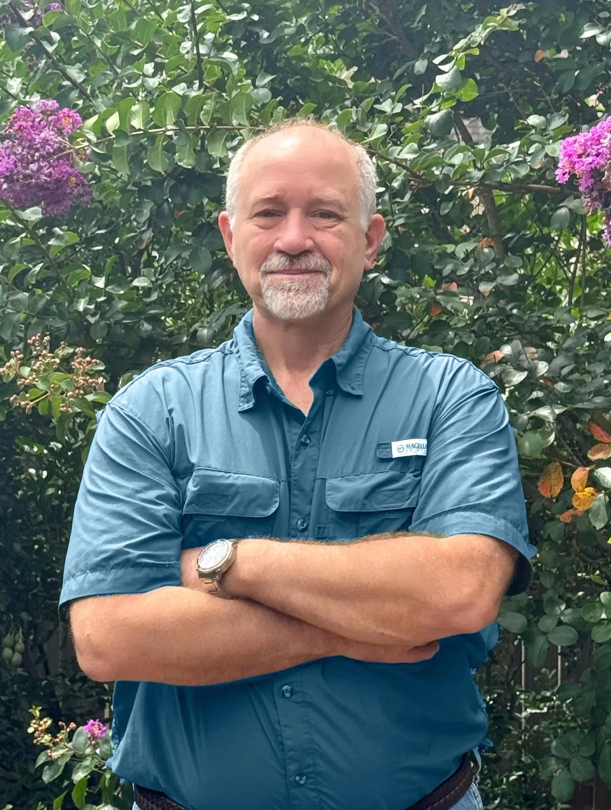 A man with a gray beard and mustache, wearing a blue short-sleeved shirt and a wristwatch, standing in front of green leafy bushes with purple flowers, with his arms crossed.