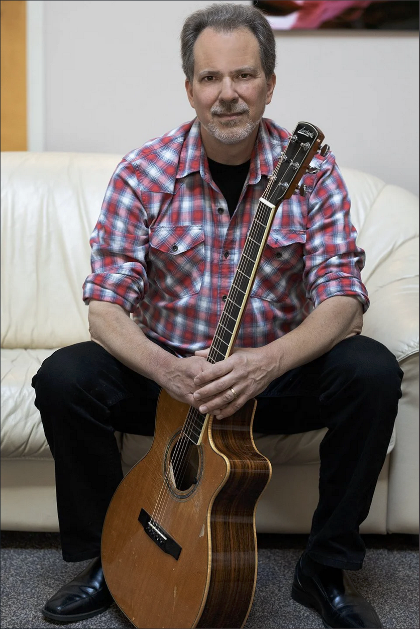 A middle-aged man with a beard, wearing a red plaid shirt, sitting on a cream-colored sofa, holding an acoustic guitar.