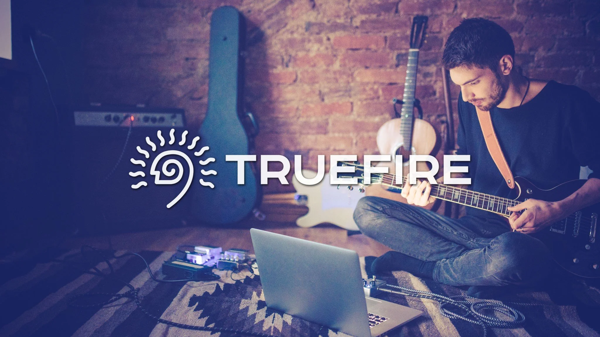 A man playing an acoustic guitar in a room with a brick wall background, surrounded by musical equipment and a laptop, with the logo and name 'TRUEIFIRE' overlayed on the image.