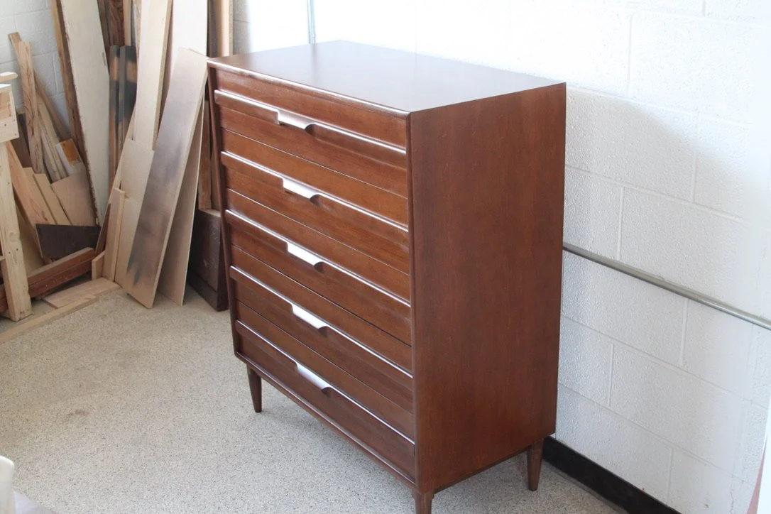 Wooden dresser with multiple drawers, standing on four tapered legs in a workshop or storage area.