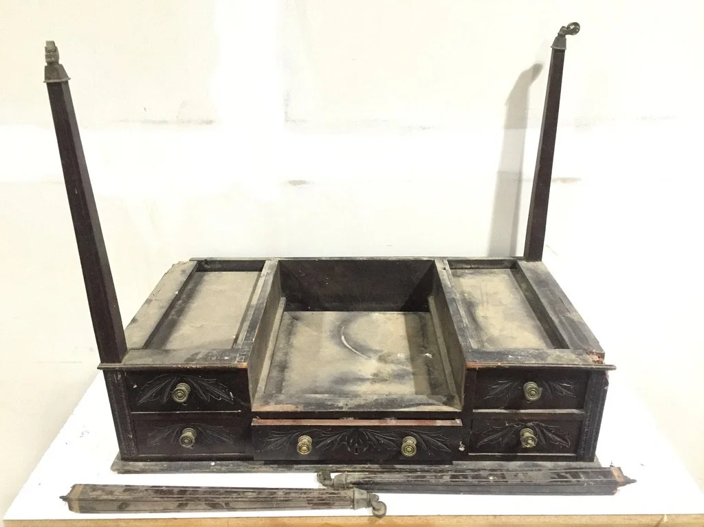 An old, black wooden desk with brass knobs, drawers, and two tall, vertical posts on each side. The desk is missing its top surface and drawers.