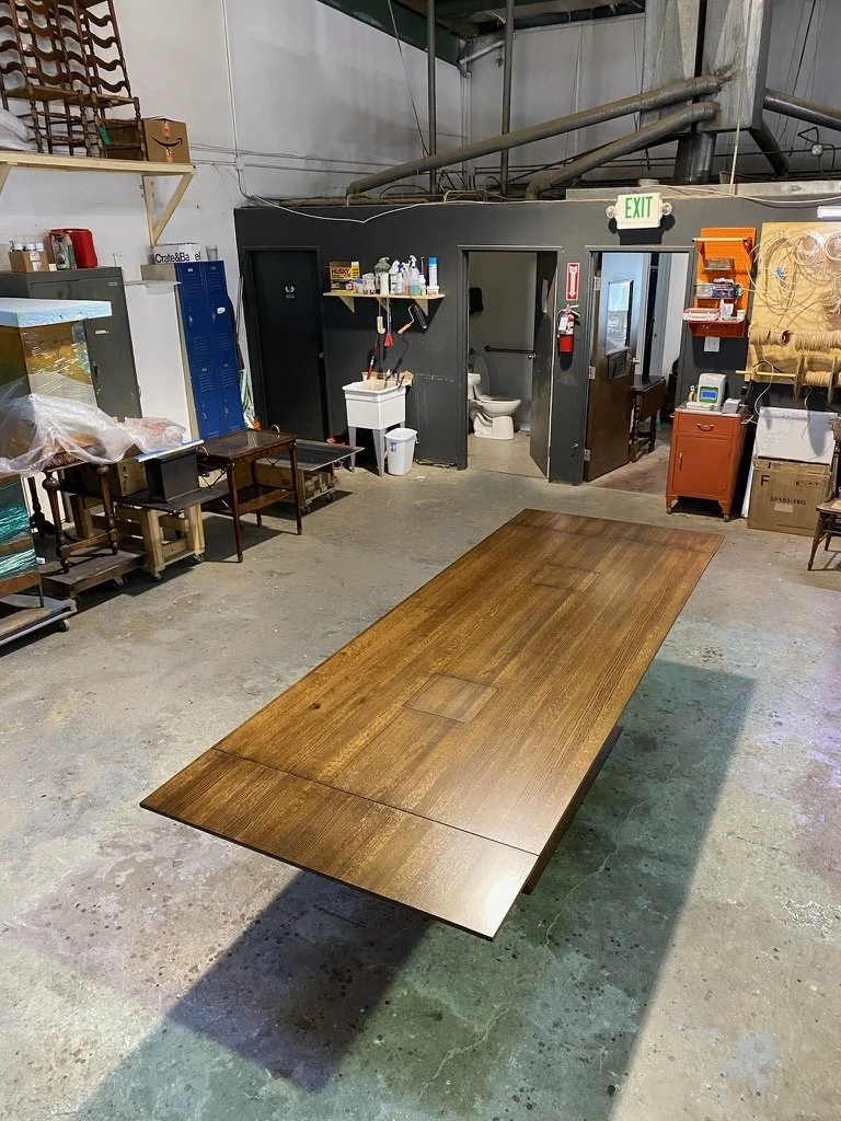 A large wooden table with a fold-out leaf in a warehouse or workshop setting.