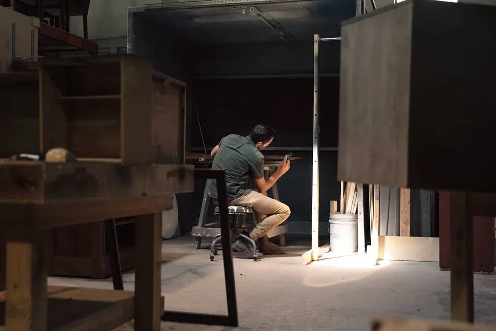 A man sitting on a stool in a woodworking or carpentry workshop, working on a piece of wood. The workshop is filled with wooden furniture and materials, with a bucket and pieces of wood visible in the background.
