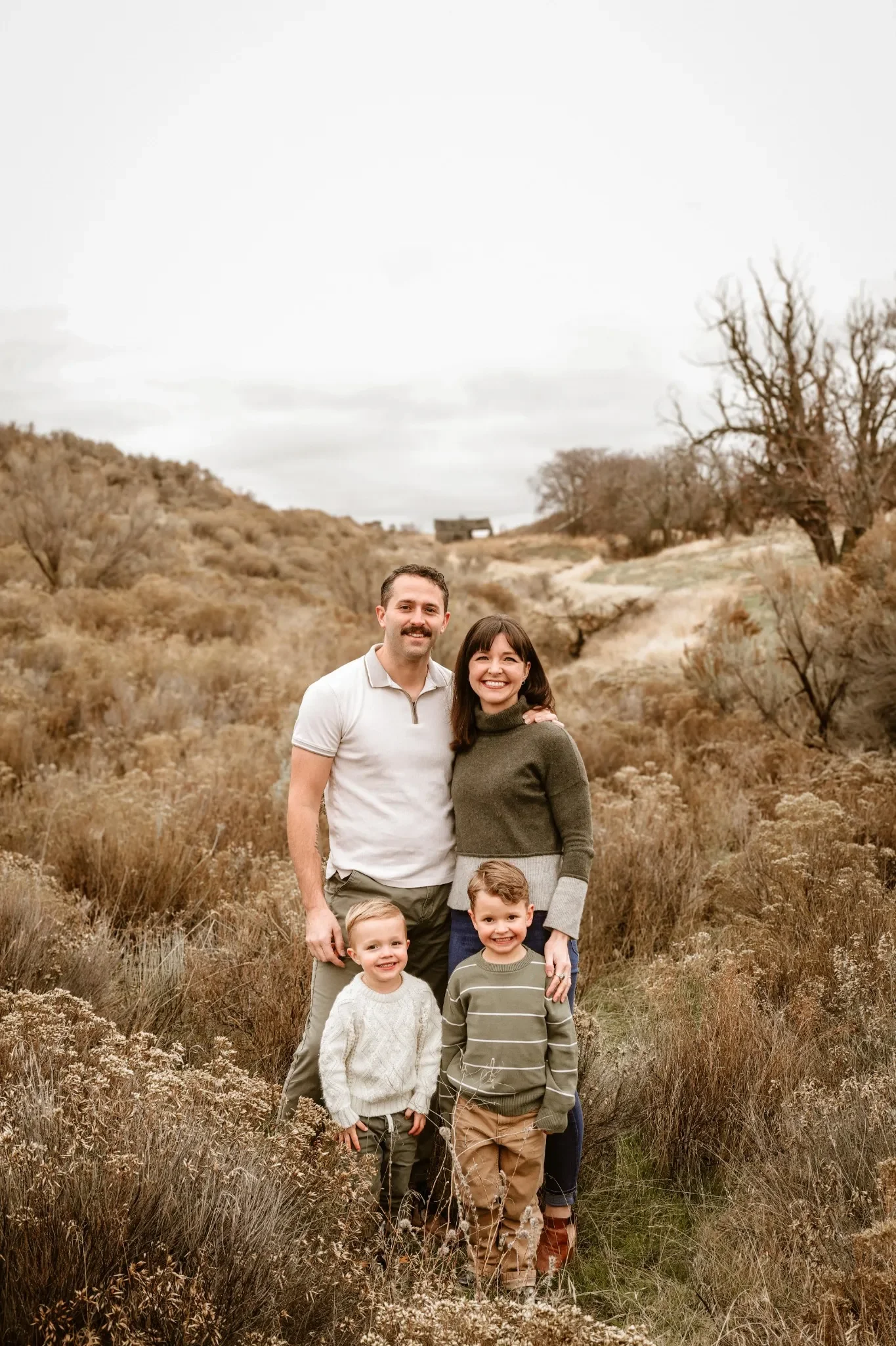 A happy family of six, including two adults and four children, stands outdoors in a dry, mountainous landscape with brown bushes and sparse trees on a cloudy day.