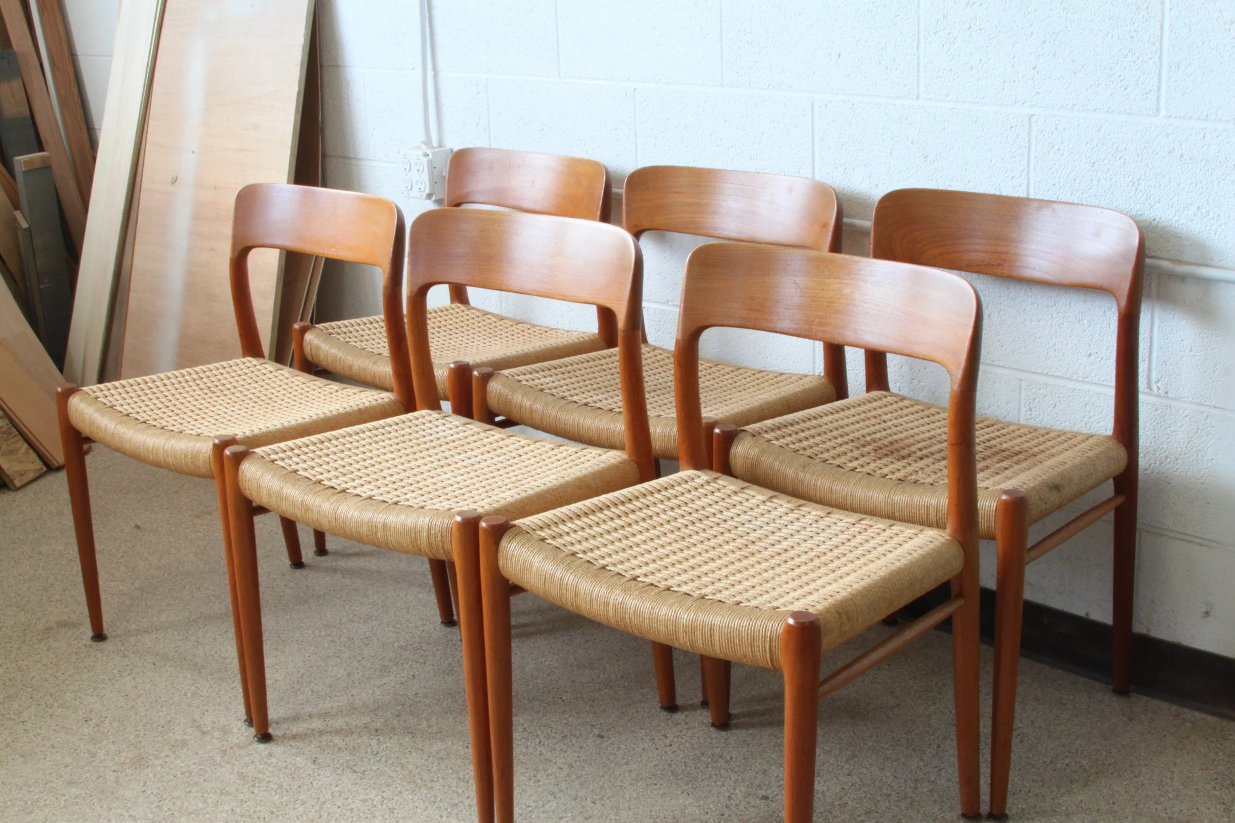A row of six wooden chairs with woven seats arranged against a white cinder block wall, with wooden boards leaning on the left side of the wall.