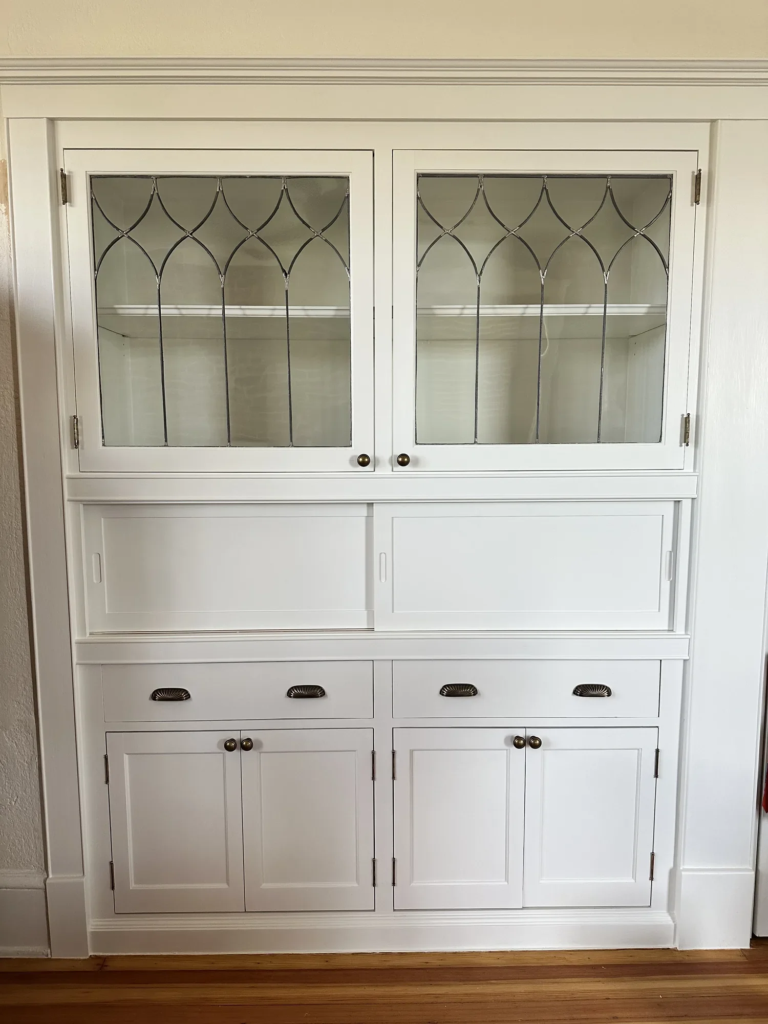 Restored white built-in cabinet with original leaded glass doors in a Gothic arch pattern, sliding panels, drawers, and lower cupboards