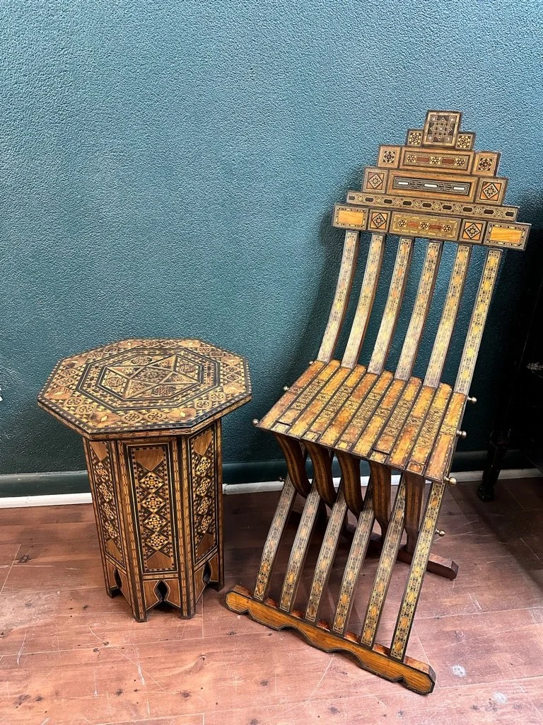 Decorative wooden side table with intricate geometric patterns next to a wooden folding chair with ornate designs, placed against a green wall on a wooden floor.