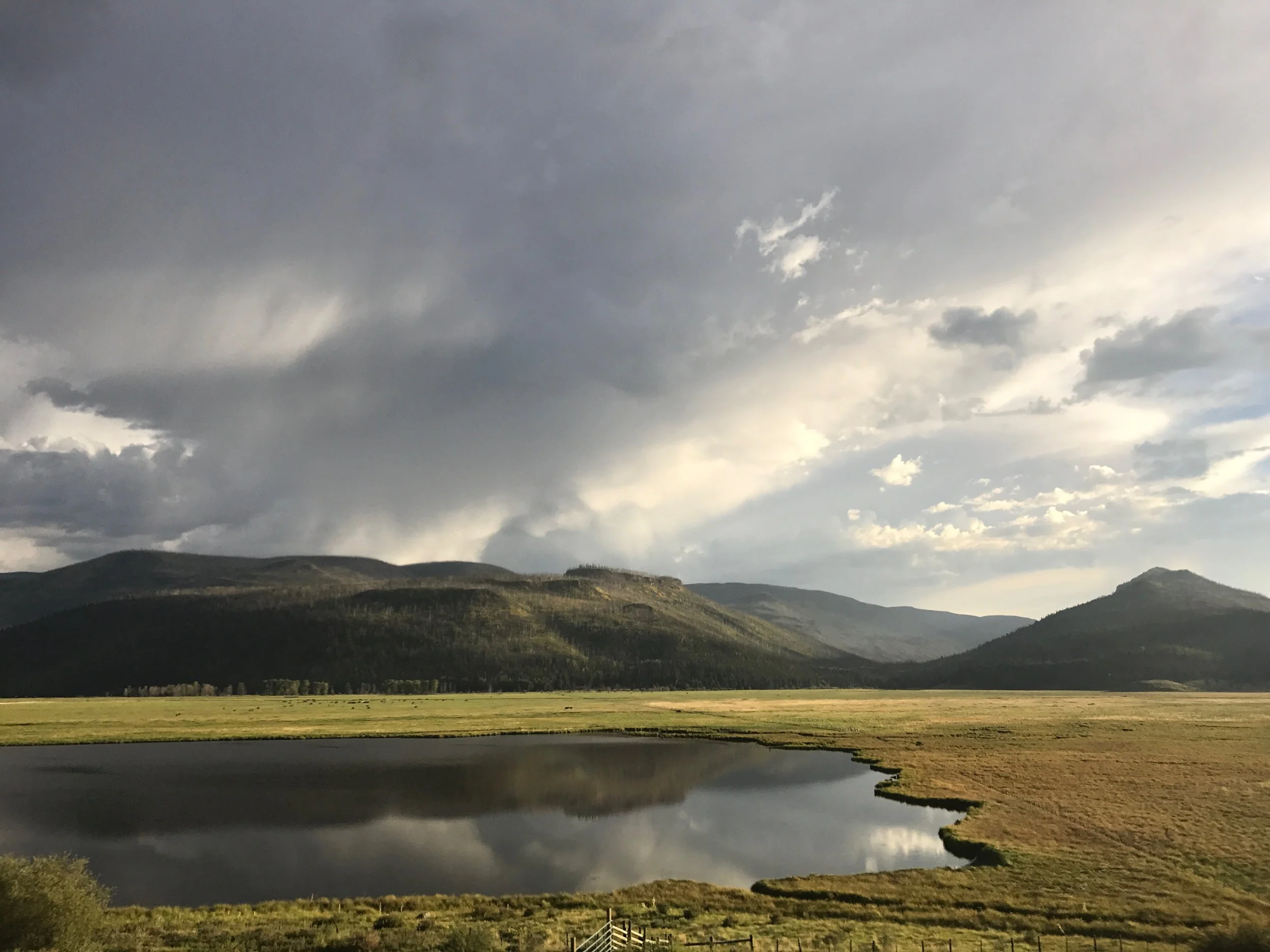A landscape with rolling green hills, a river reflecting the cloudy sky, and a fence in the foreground.