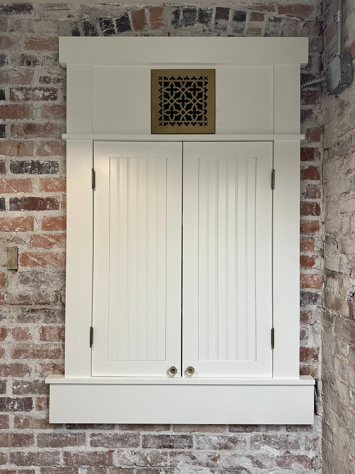 White built-in wall cabinet with beadboard doors, glass knobs, and a decorative brass grille, set against exposed brick