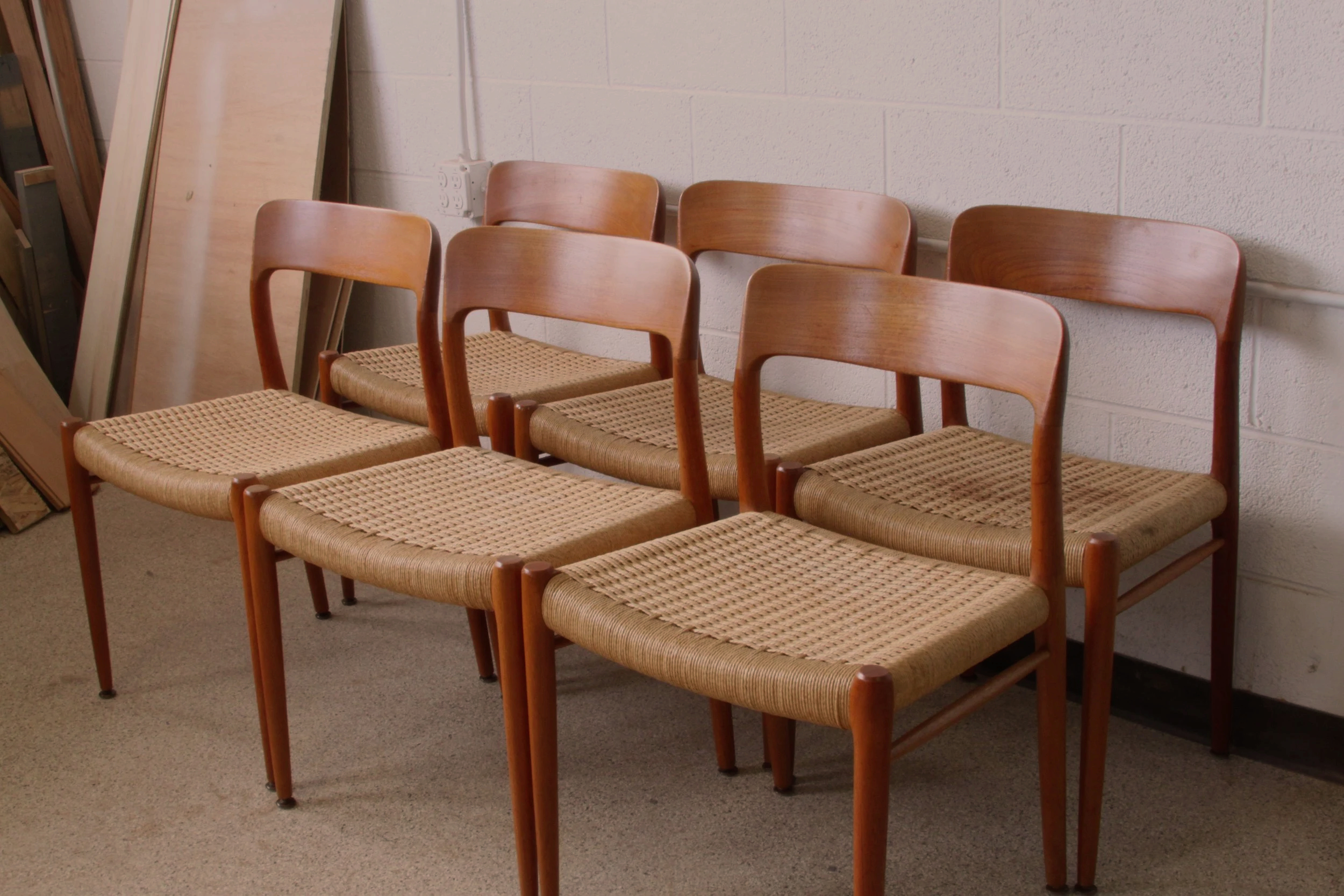 Six wooden chairs with woven seats lined up against a white brick wall in a workshop or storage room.