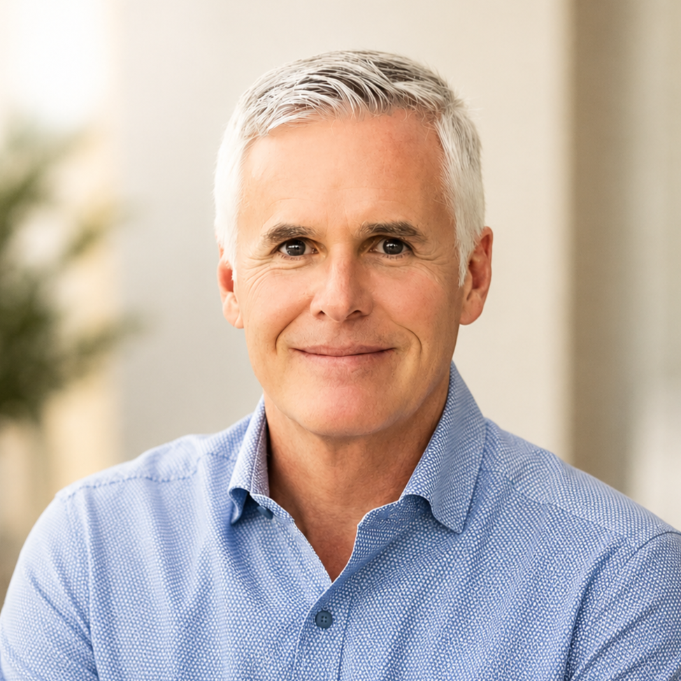 A middle-aged man with short gray hair wearing a light blue button-up shirt, smiling in an indoor setting.