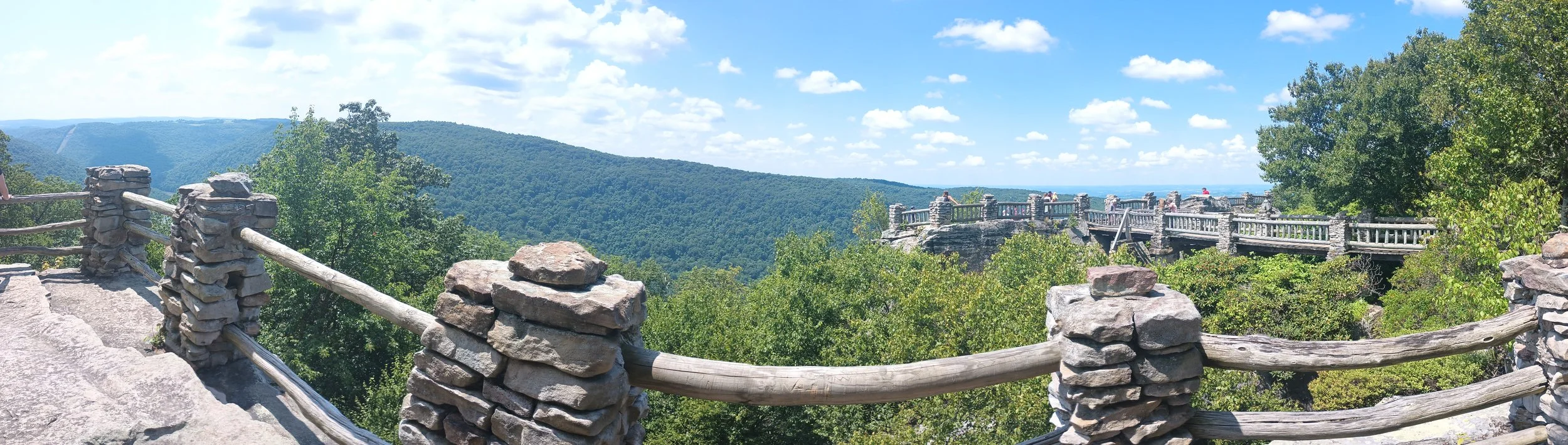 View from a stone and wooden railing overlook showing green trees, a mountain range, and a blue sky with clouds.