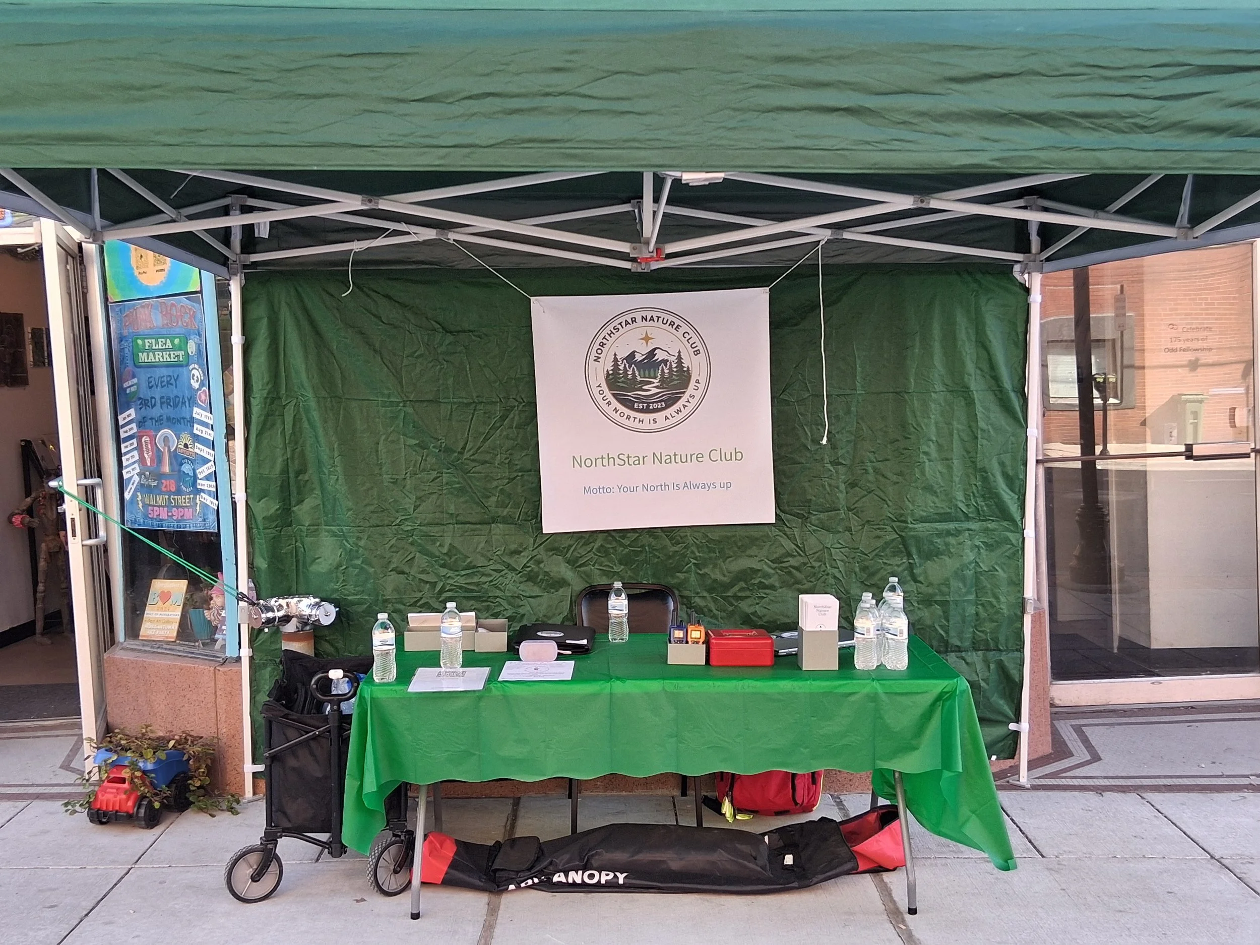 A booth at an outdoor event for NorthStar Nature Club with a green tablecloth, several water bottles, informational materials, and a banner displaying the club's logo and motto, 'Your North Is Always Up.'