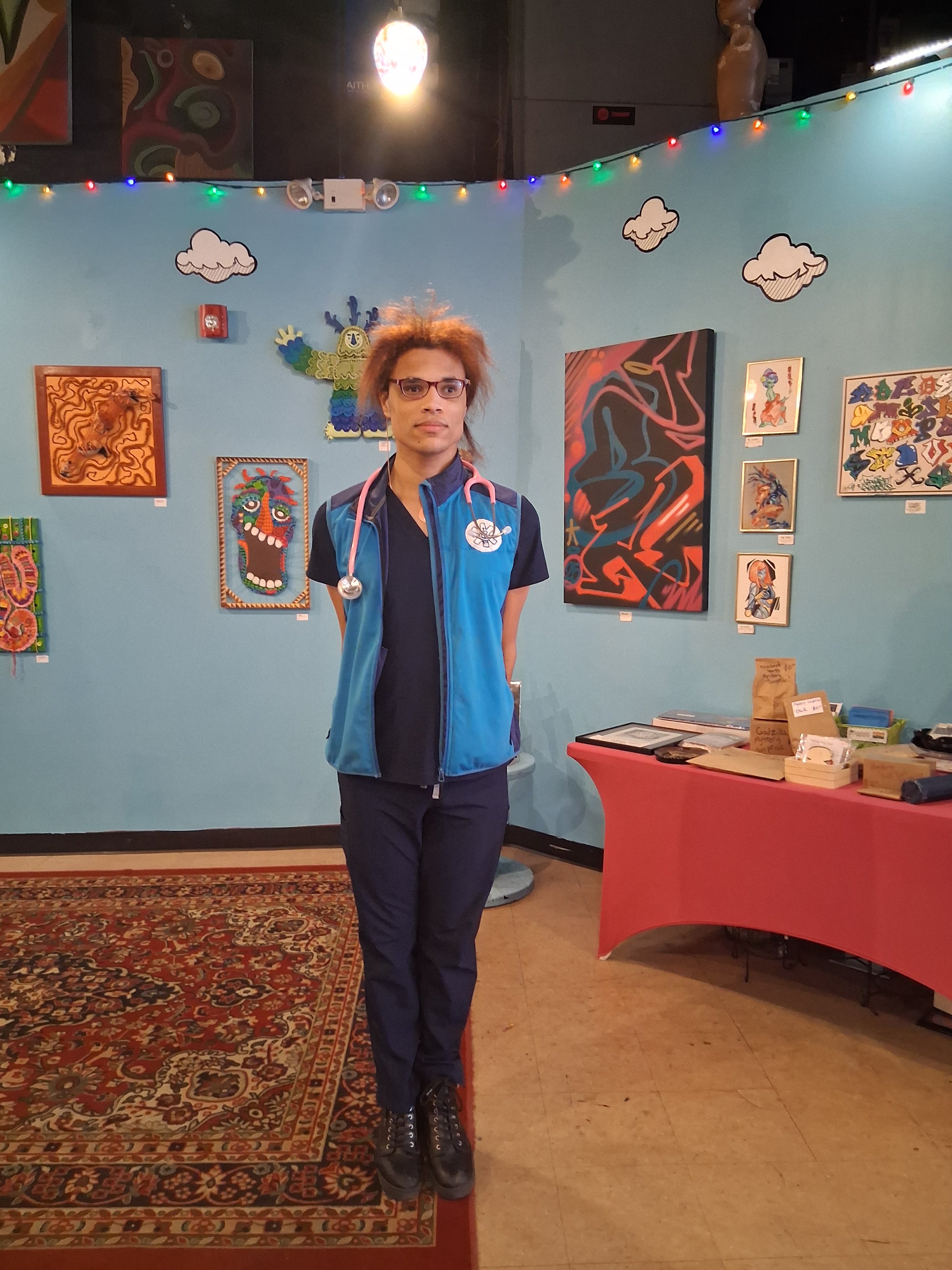 A person with curly, reddish hair, wearing glasses, a blue and black jacket, and black pants, standing in an art gallery with colorful paintings on the blue wall behind them. The gallery has a red table with items on it and string lights hanging over