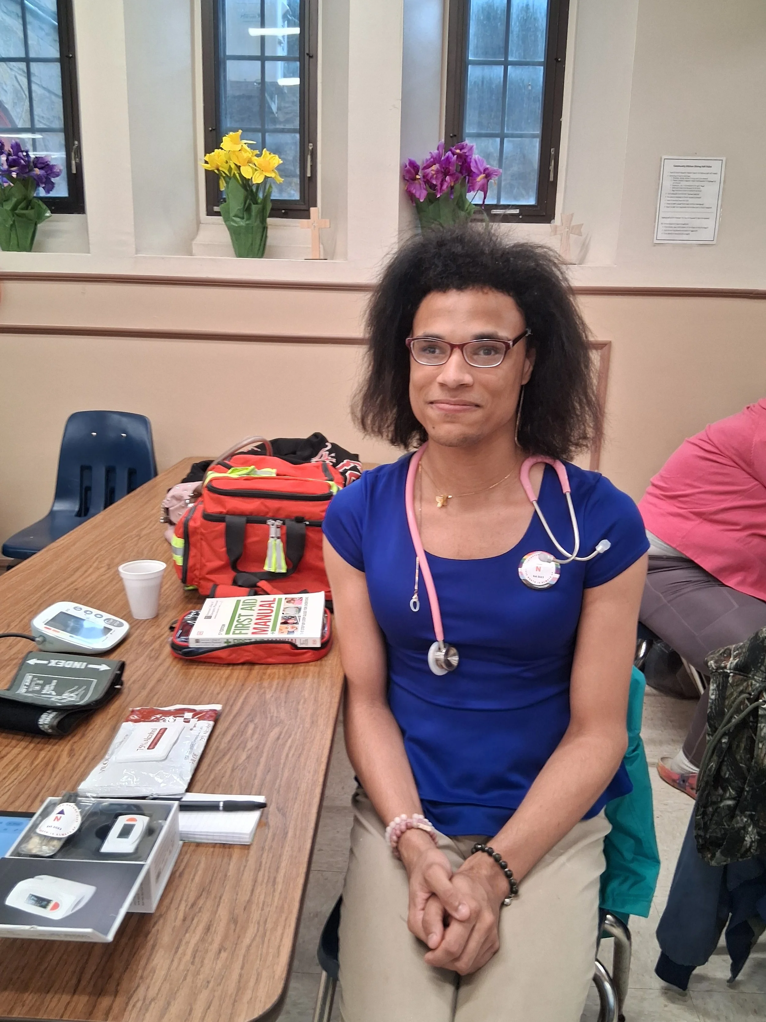 A woman with glasses and a stethoscope sitting at a table. The table has medical supplies, a clipboard, and a magazine. There are colorful flower bouquets in the background by the window.