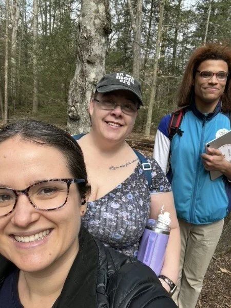Three women taking a selfie outdoors in a wooded area, smiling, wearing glasses, and casual clothes.