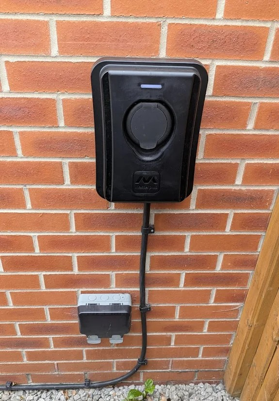 Electric vehicle charging station mounted on a brick wall with a connected cable and a gray junction box below.