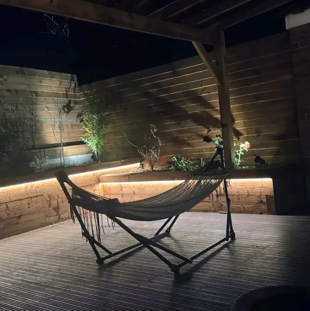 A hammock on a screened porch or patio with wood flooring and wall, plants, and outdoor lighting.