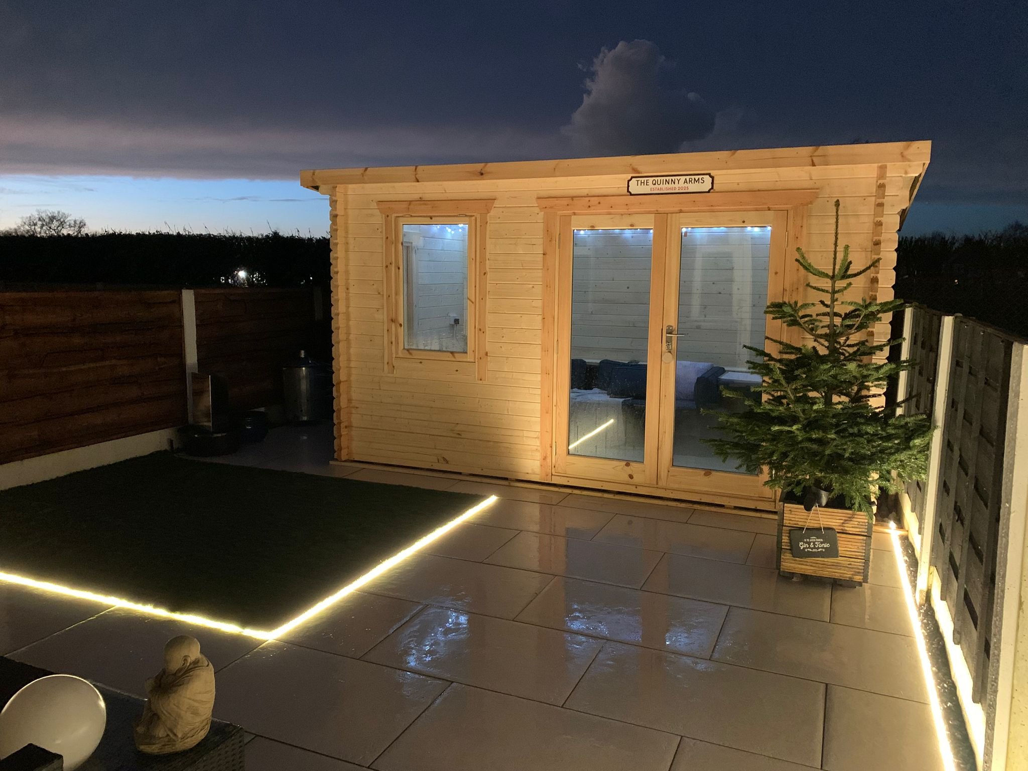 A small wooden building with a sign reading 'The Quinny Arms, Established 2023' features large windows with blue lights inside. The outdoor area has modern LED strip lighting along the floor and a small Christmas tree placed outside. The sky is dark with clouds, indicating dusk or evening.