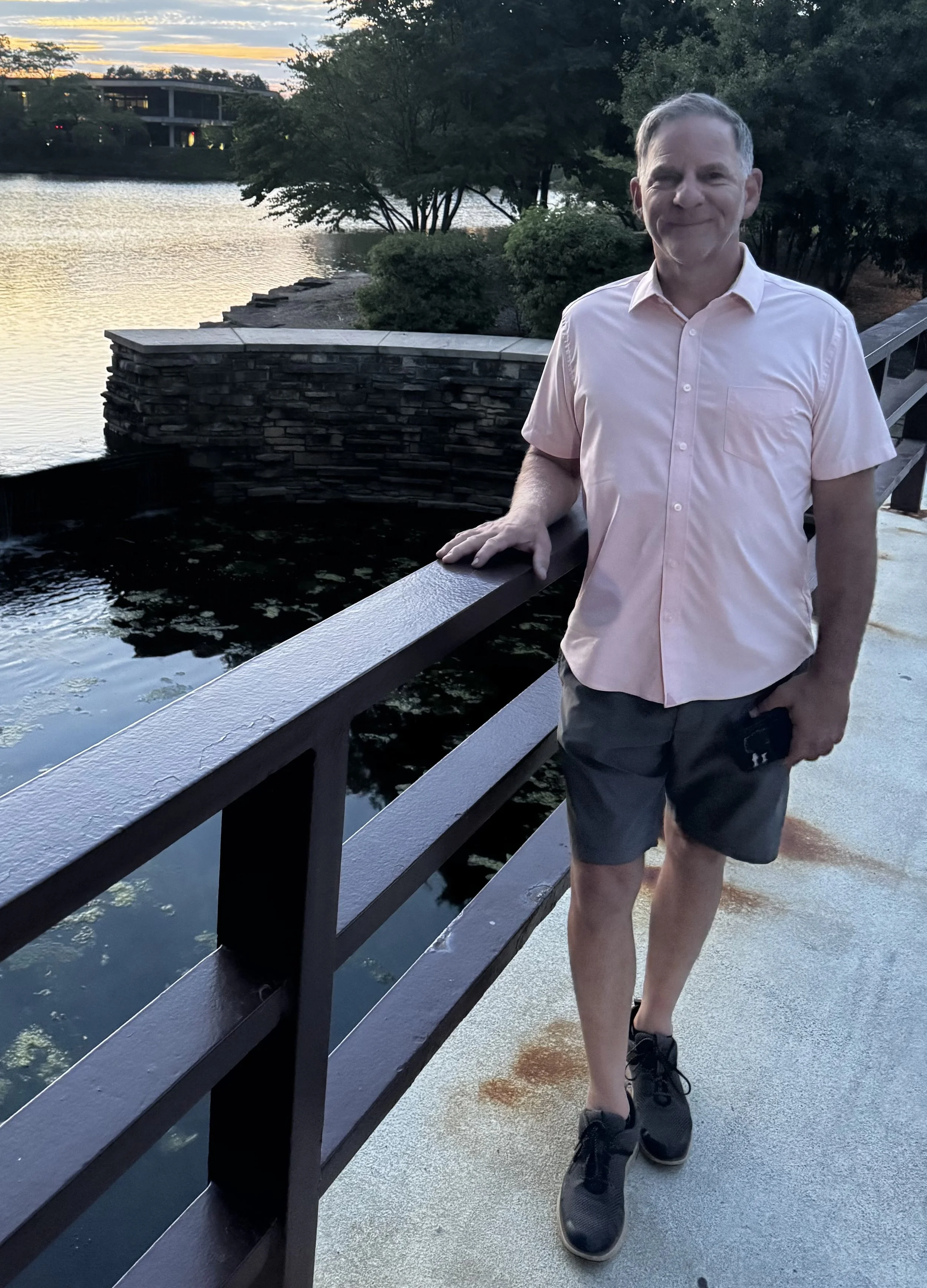 A man wearing a light pink shirt and black shorts walking on a paved path by a body of water with trees and a sunset in the background.