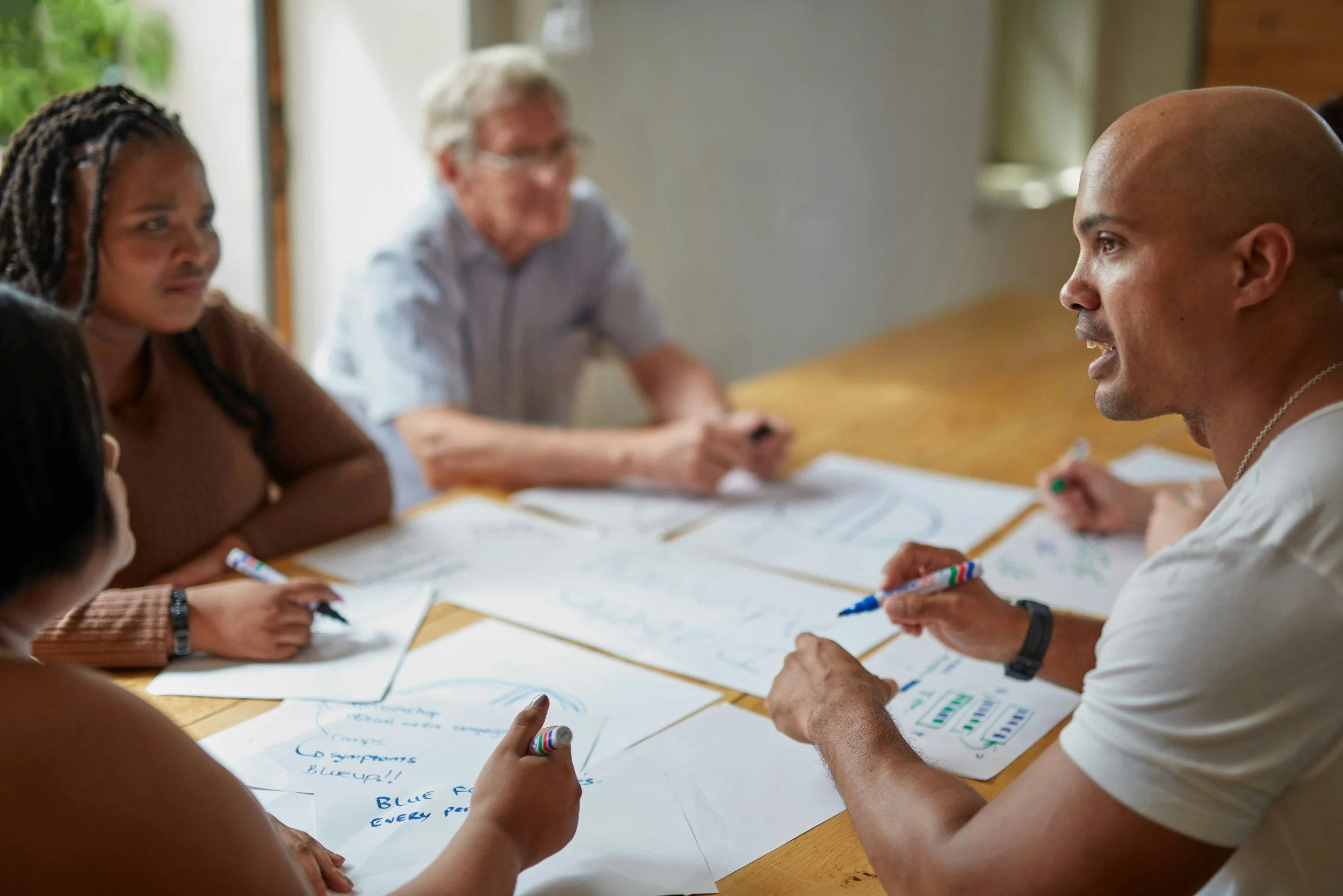 Group of diverse people sitting around a table, engaging in a discussion or brainstorming session with written notes and drawings on paper.