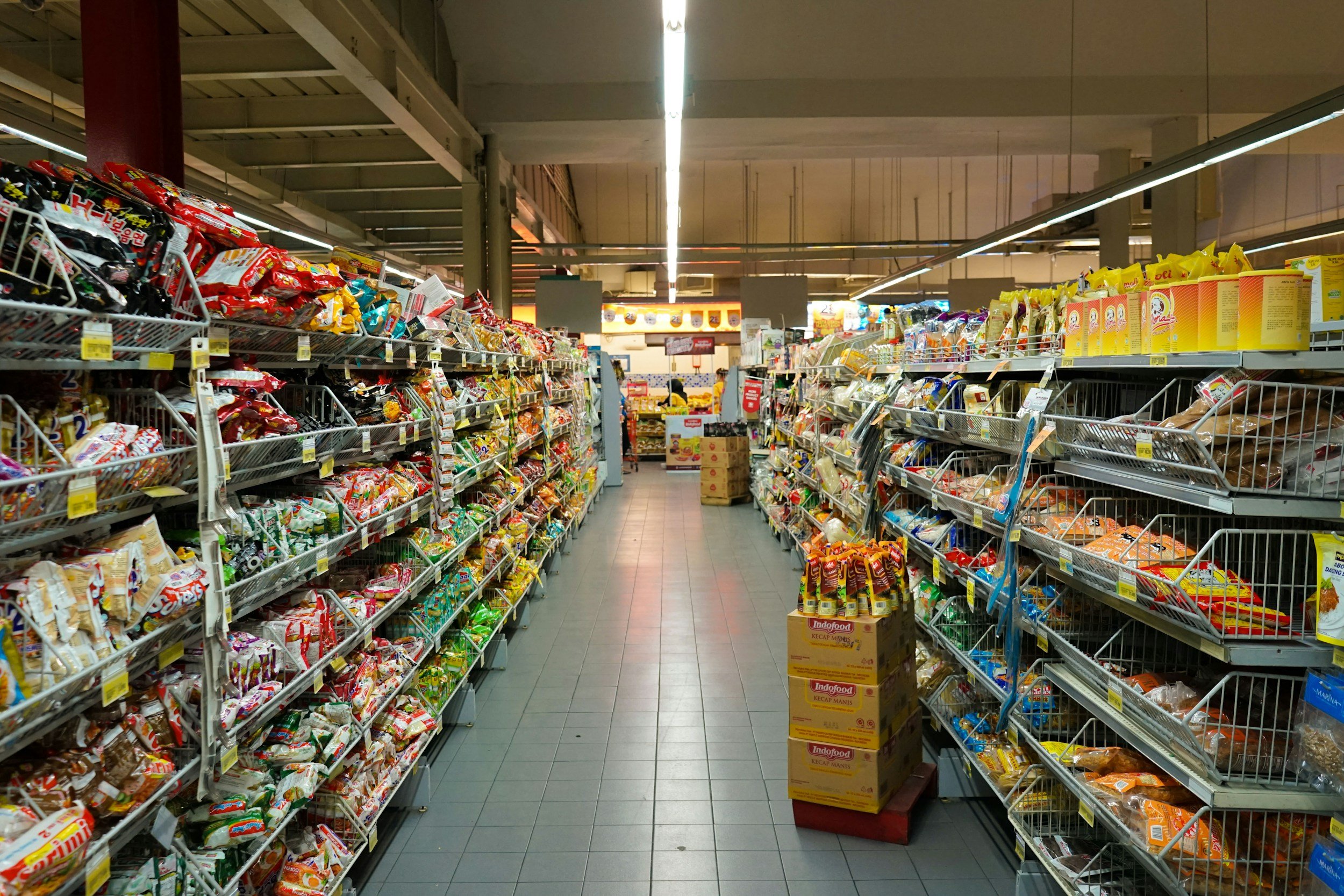 A supermarket aisle with shelves on both sides filled with snack packages, with a few boxes and shopping carts in the aisle, and bright ceiling lights overhead.