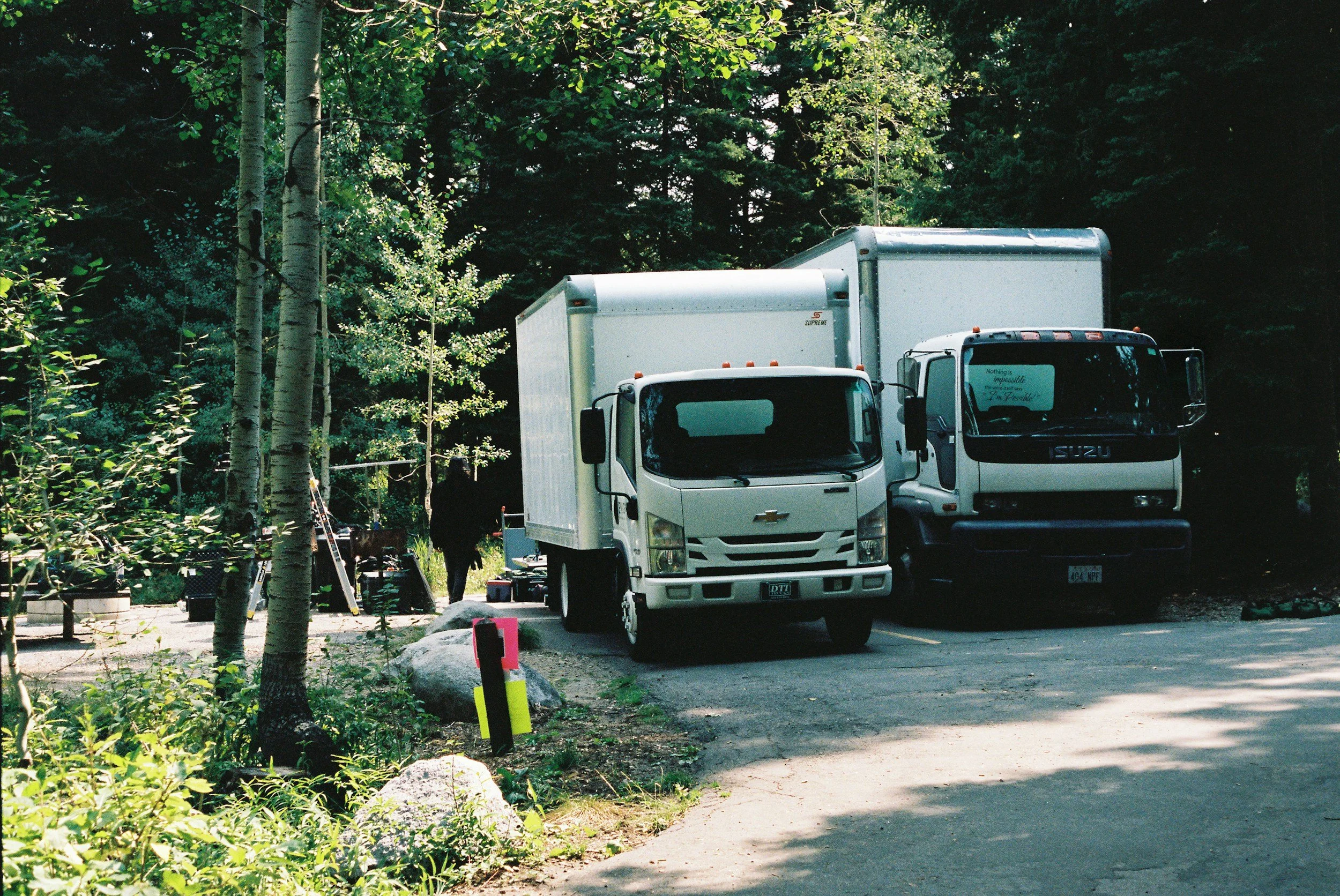 A parking area in a wooded area with several trees and two white trucks parked side by side, and a person standing near the trucks.
