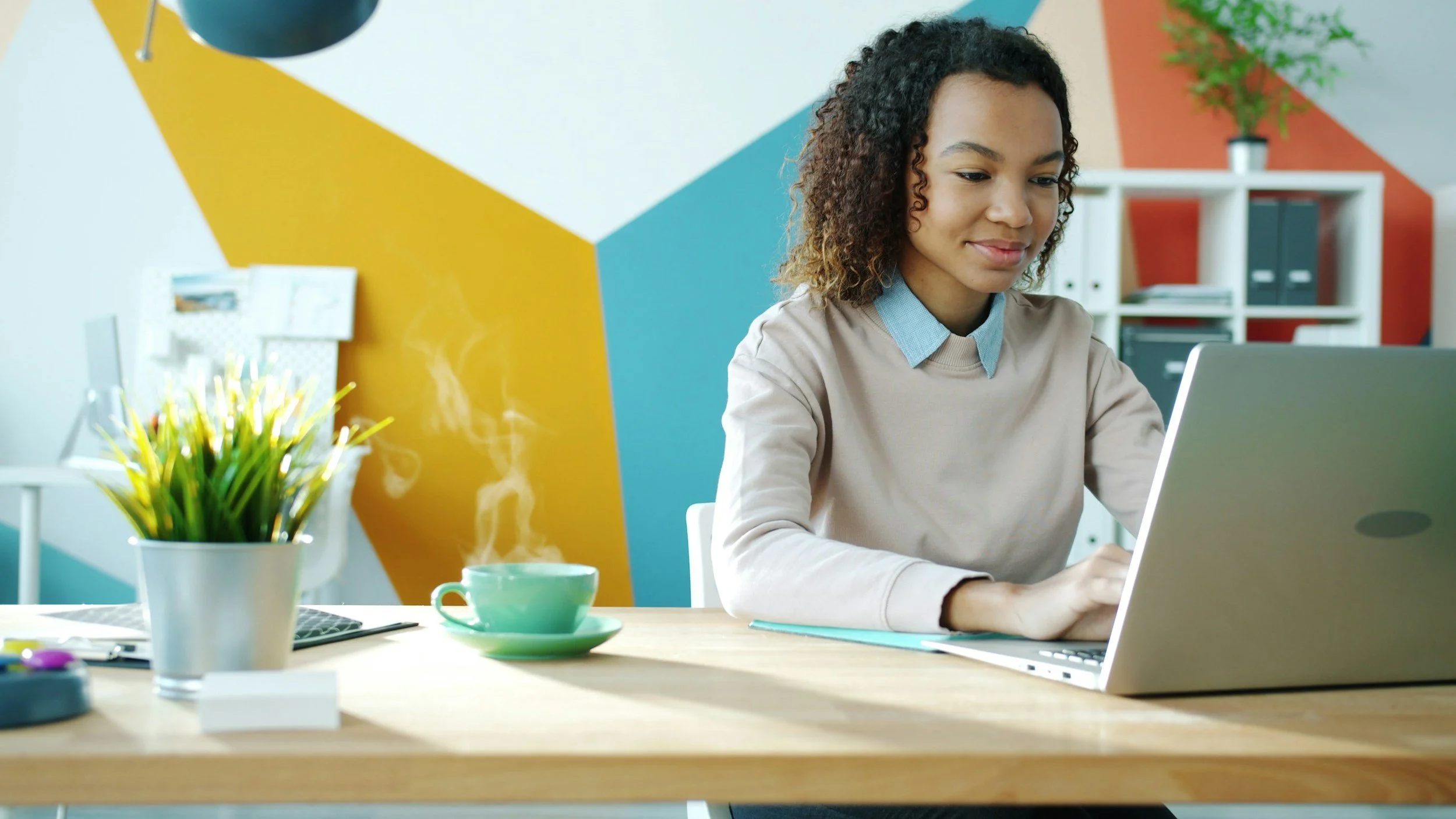 A young woman with curly hair working on a laptop at a modern, colorful office desk with plants and a yellow, blue, and orange wall pattern.