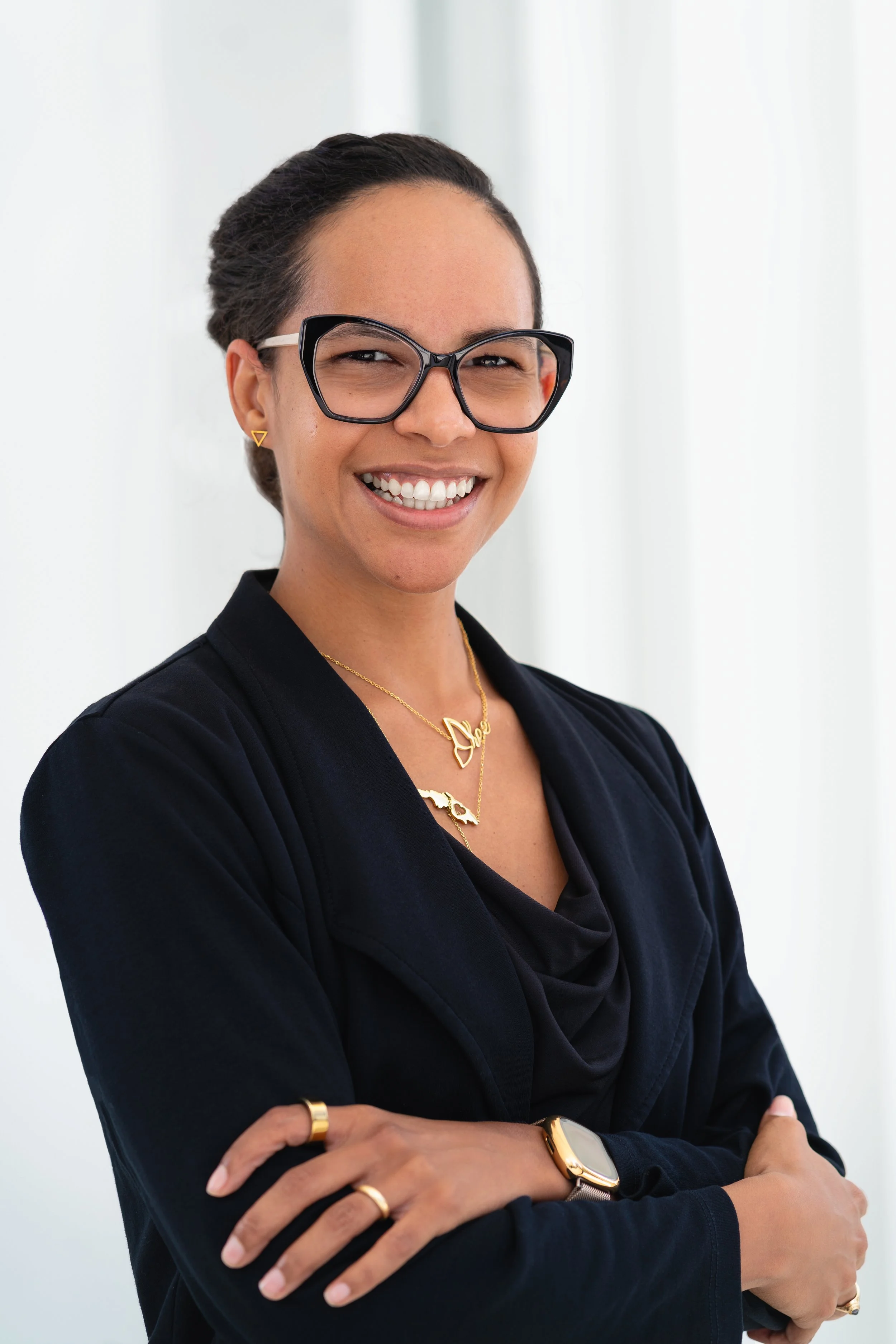 A woman with dark hair styled in an updo, wearing black-framed glasses, gold jewelry, a black blazer, and a watch, smiling with her arms crossed in front of a white background.