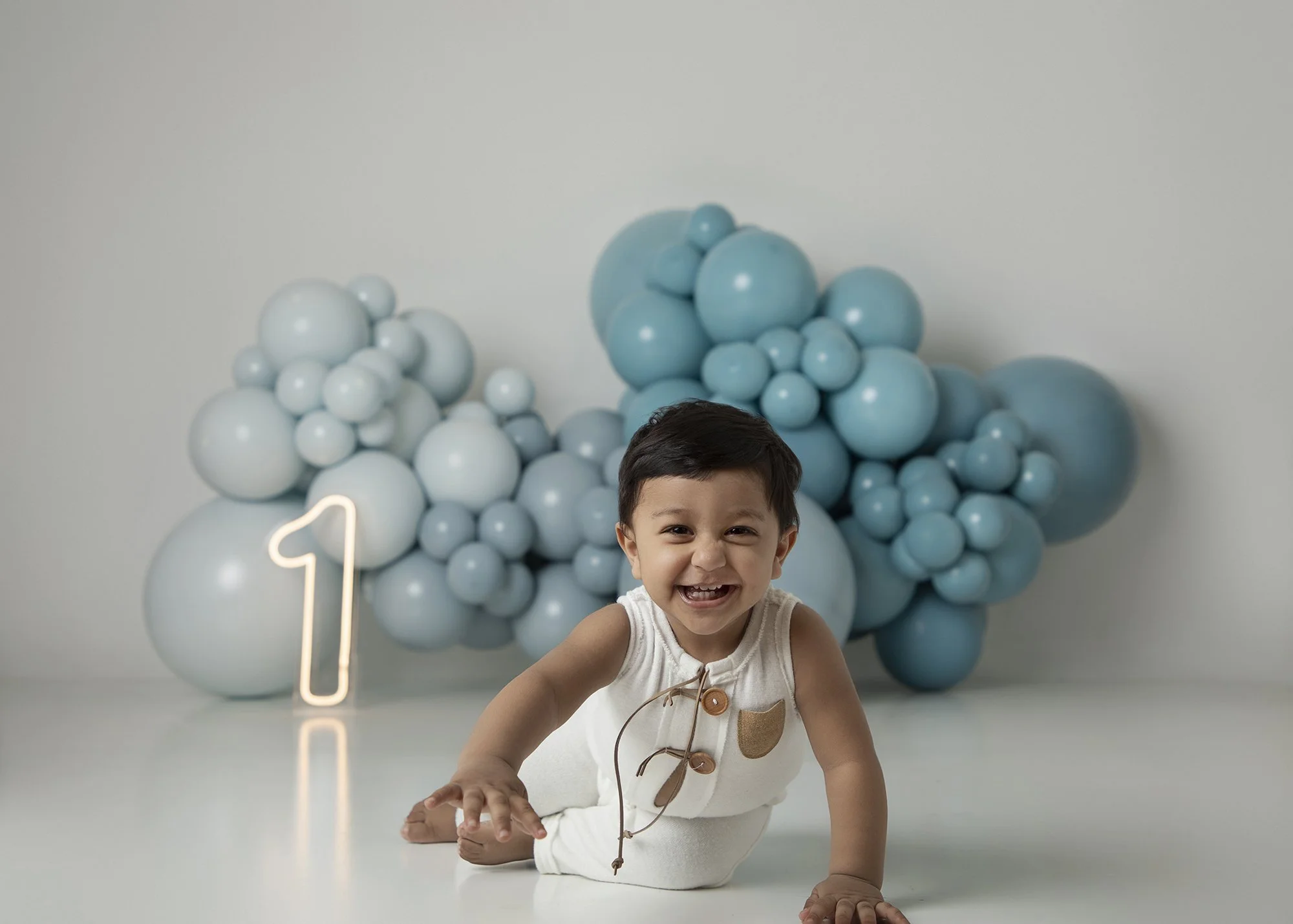 A young boy with dark hair crawling on a white surface, smiling at the camera, in front of a backdrop decorated with large blue and white balloons and a neon number one sign, indicating a first birthday celebration.